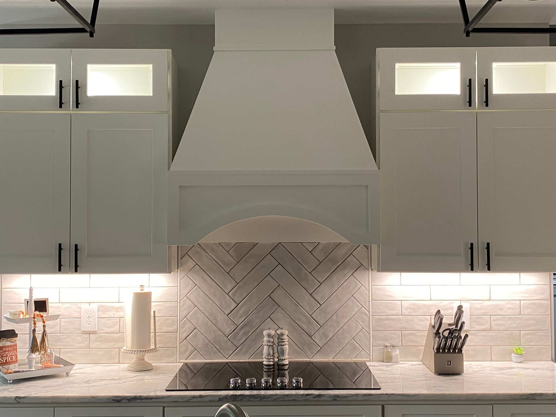 Modern kitchen with white cabinets, herringbone backsplash, stainless range hood, and under-cabinet lighting