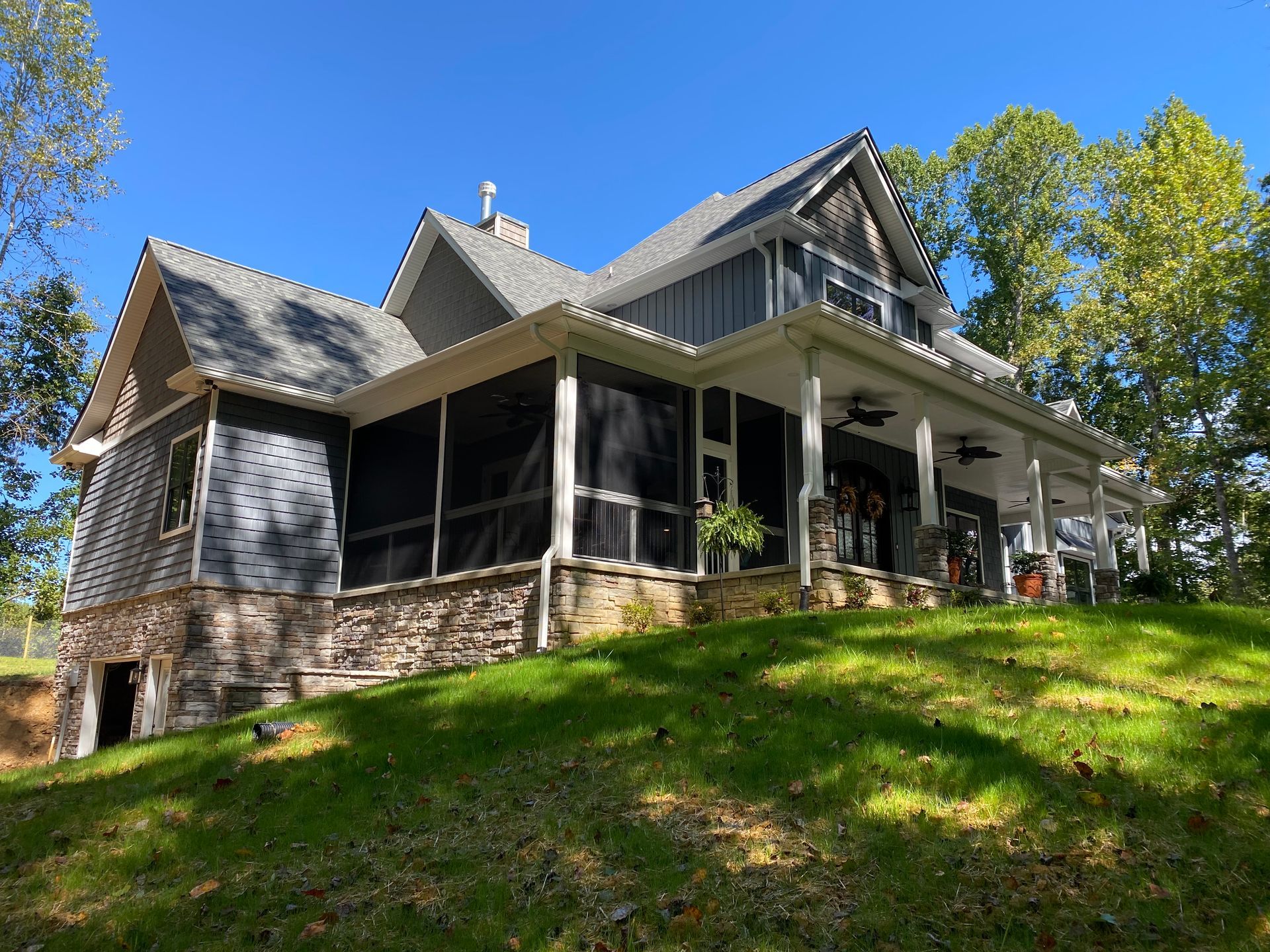 Large gray house with a wraparound porch on a sunny wooded hillside
