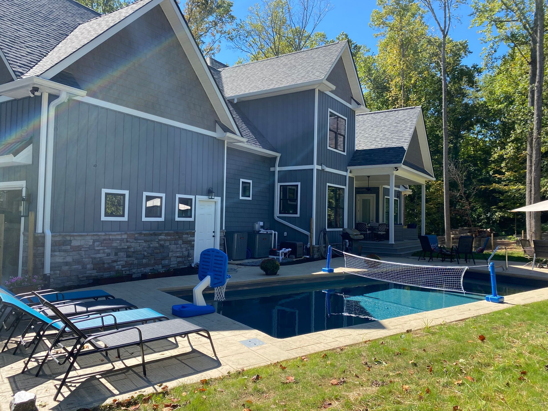Backyard pool beside a gray house with lounge chairs, a covered patio, and tall trees.