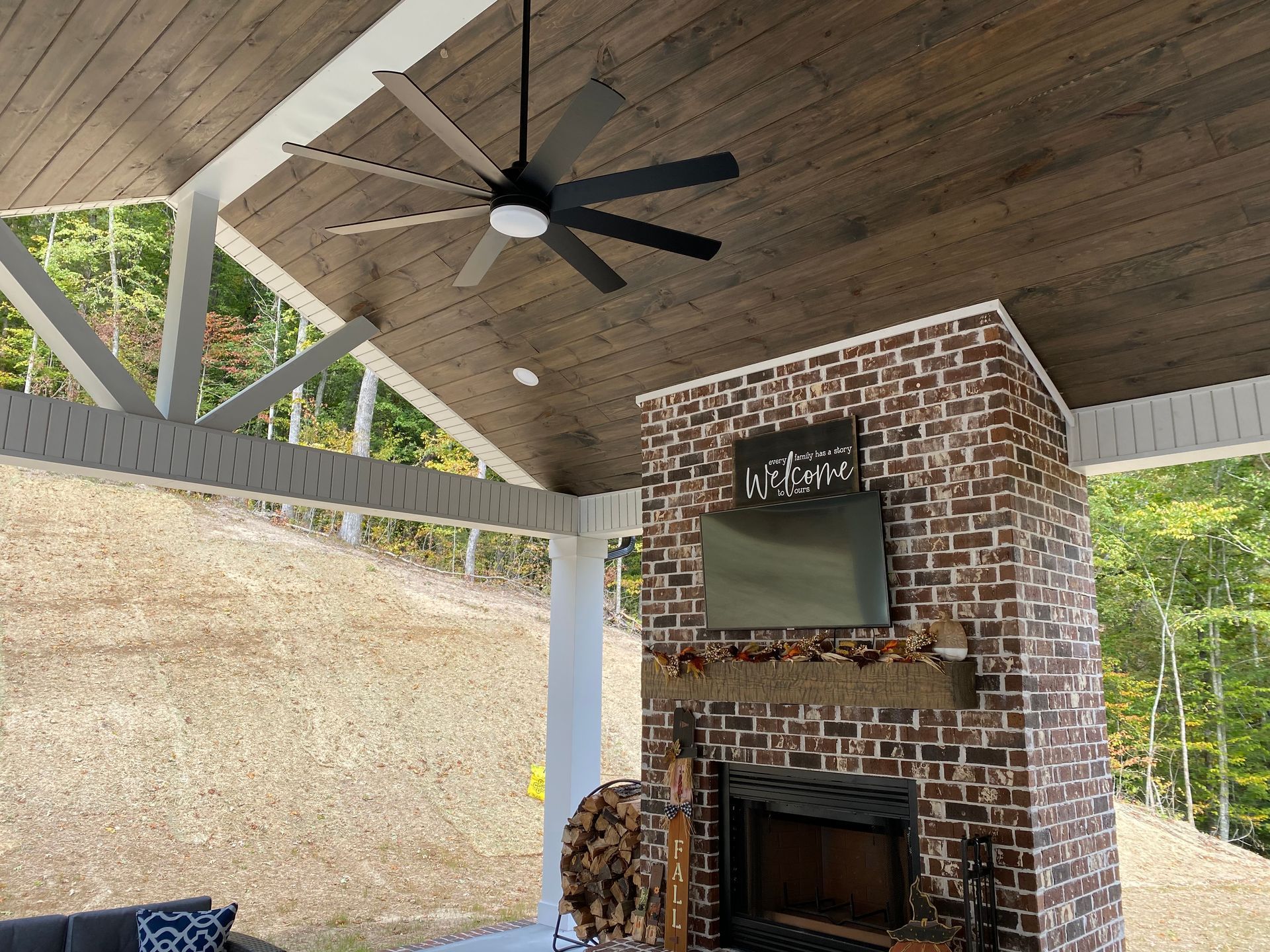 Covered outdoor fireplace under a wooden pavilion with a ceiling fan and TV mounted on a brick chimney