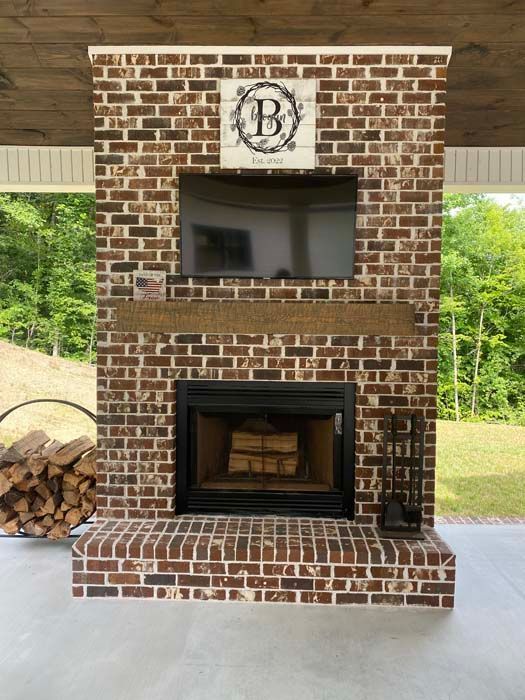 Brick outdoor fireplace under a covered patio with a monogram sign above and stacked firewood beside it