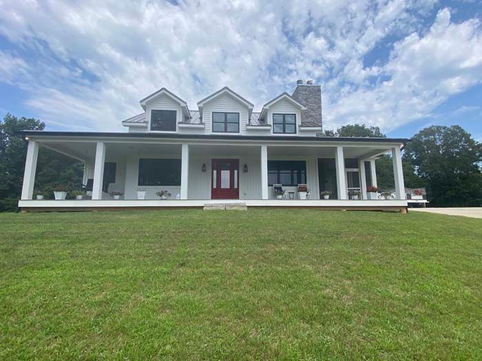Large white farmhouse with a wraparound porch and red front door on a grassy lawn