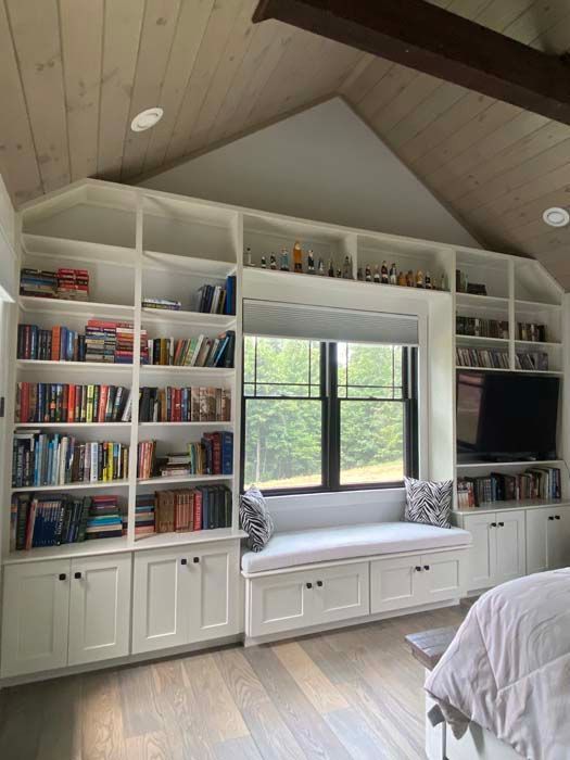 Built-in white bookshelves framing a window seat in a cozy attic bedroom with wood-paneled vaulted ceiling.