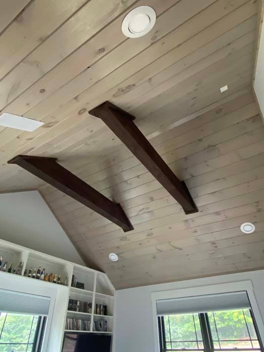 Ceiling with light wood planks, dark beams, and recessed lights above a bright room with windows.