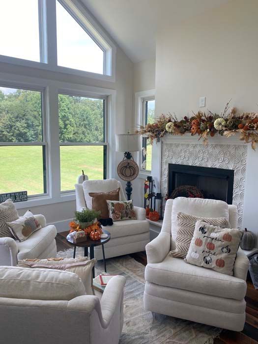 Bright living room with large windows, white chairs, and a fireplace decorated with autumn garland.