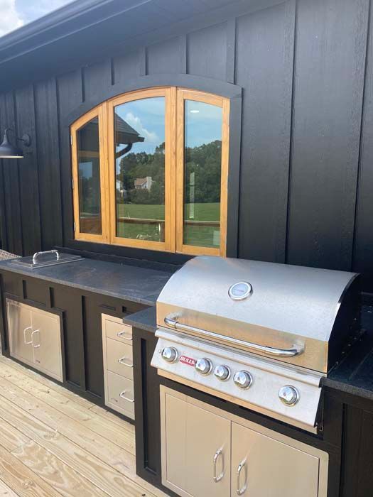 Outdoor kitchen with black cabinets, wood-framed window, and stainless steel grill on a deck.