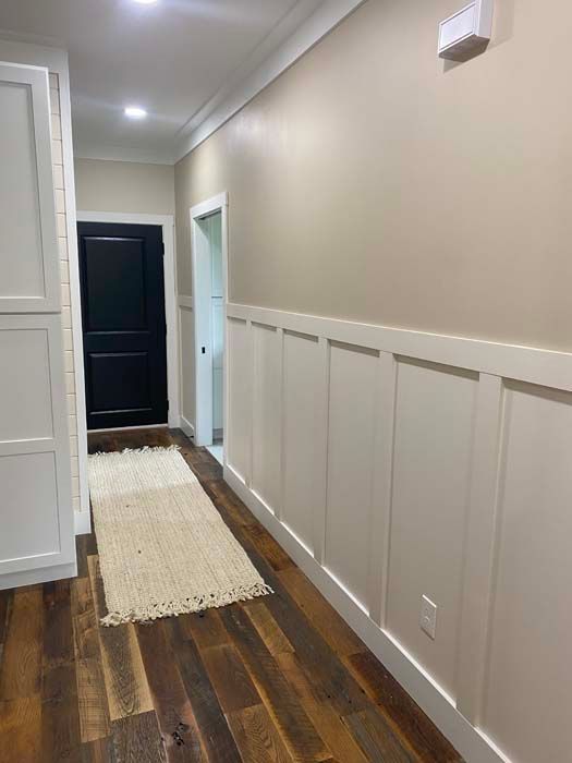 Narrow hallway with white paneled walls, dark wood floor, beige rug, and black door at the end