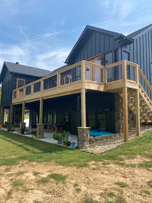 Modern dark-sided house with a wooden deck, stone pillars, and a blue hot tub under a partly cloudy sky