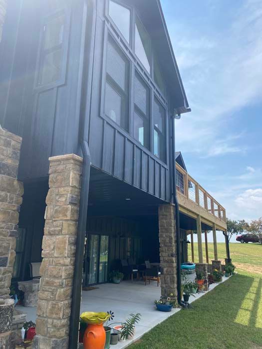 Modern house exterior with dark siding, stone columns, covered patio, and green lawn on a sunny day
