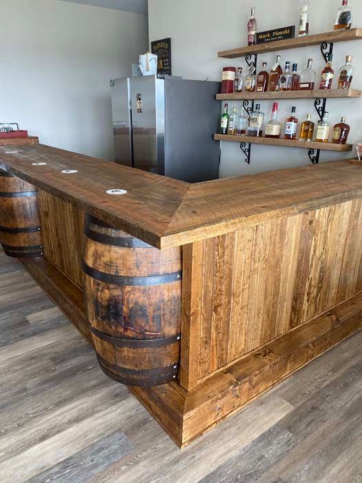 Rustic wood bar with barrel ends, shelves of liquor bottles, and a stainless-steel refrigerator in a modern room