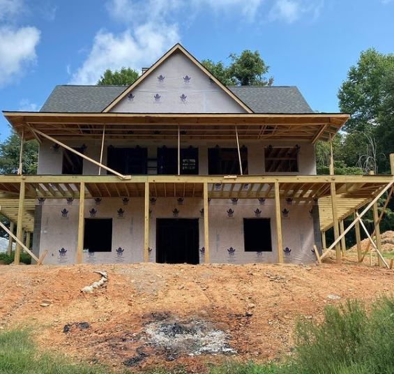 Two-story house under construction with exposed framing, white siding, and a dirt front yard under a blue sky