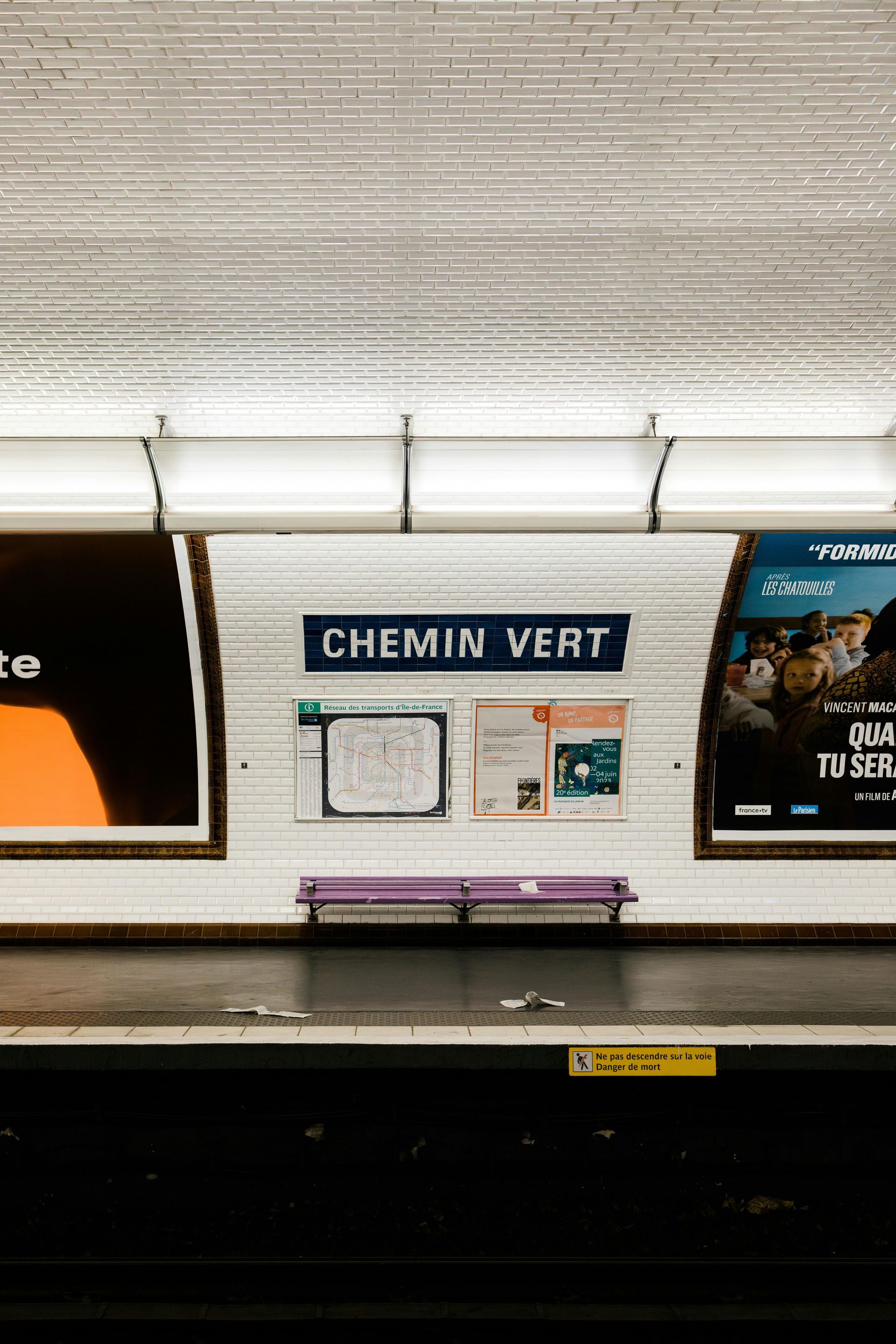 Quai de la station de métro Chemin Vert avec panneau, affiches, banc violet et éclairage au plafond.