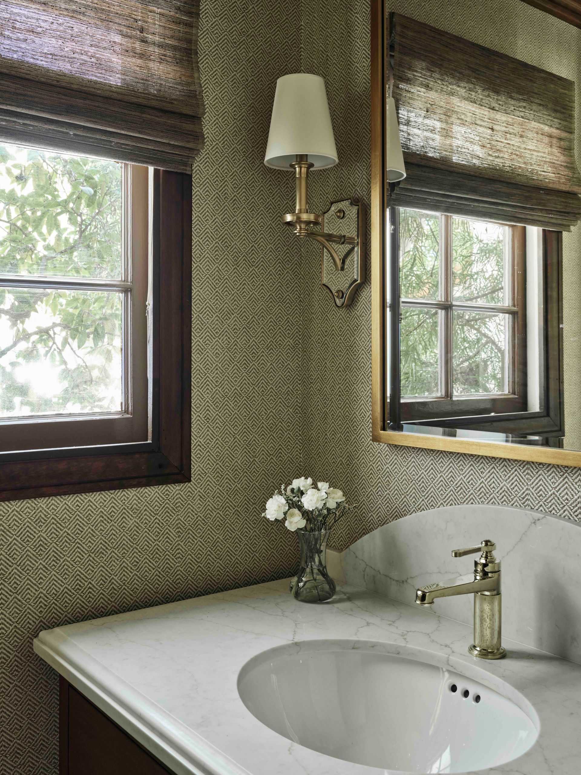 A bathroom vanity with a white marble countertop, gold faucet, wall-mounted sconce, and textured wallpaper.