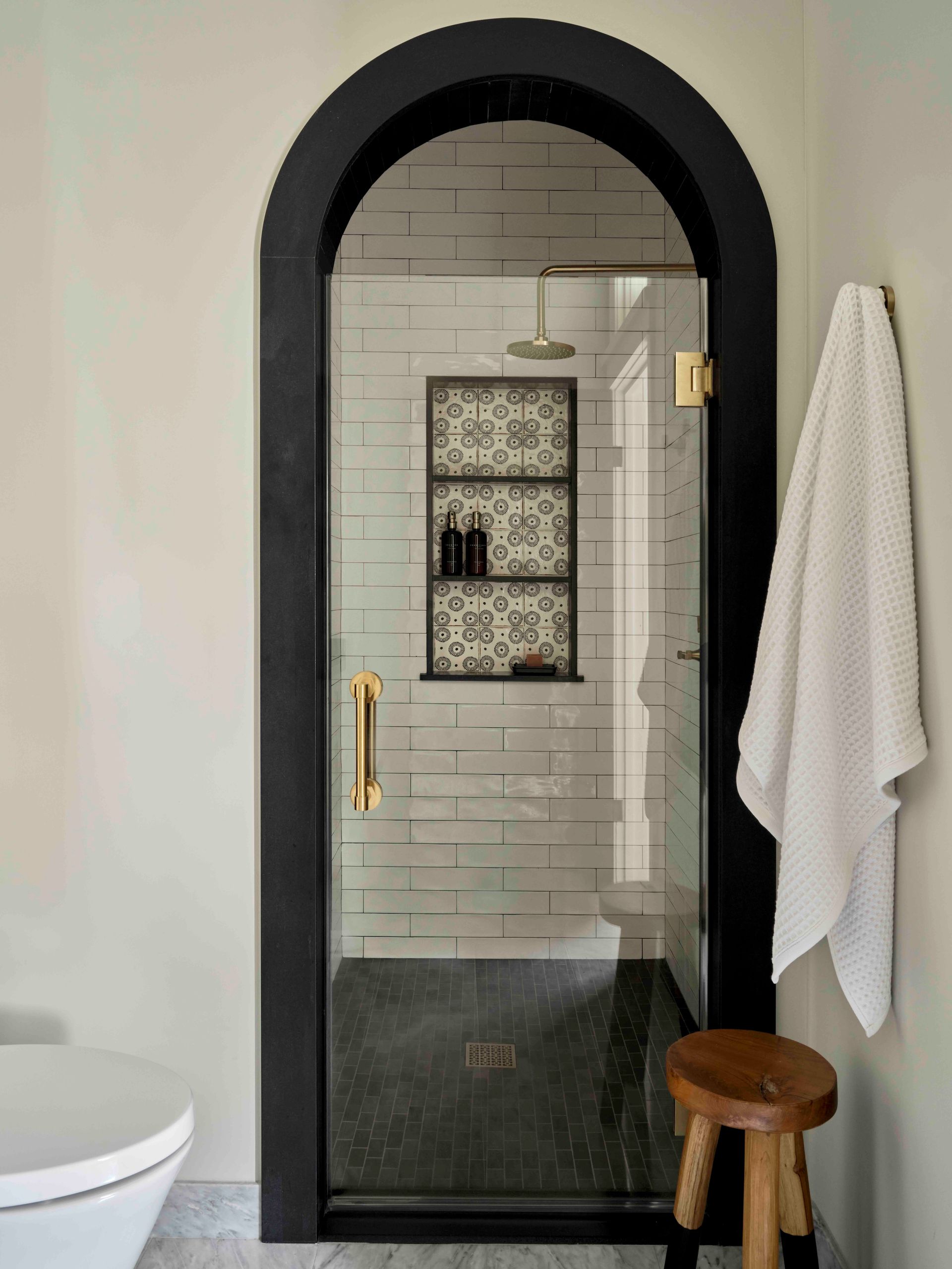 An arched shower door with a black frame, gold handle, frosted glass, and patterned tile niche, next to a white towel.