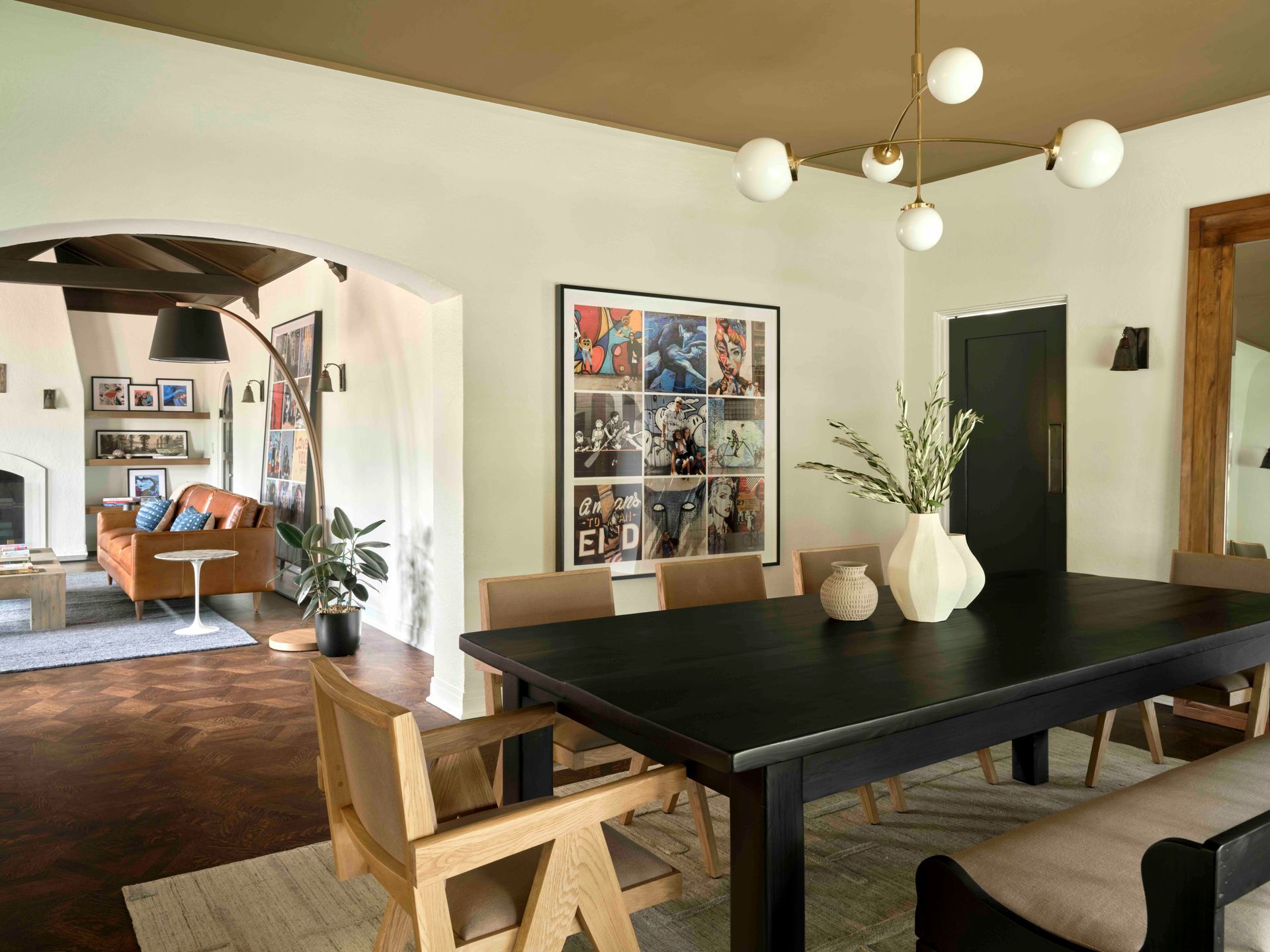 A black dining table sits in a light-walled room with a brown ceiling, a modern light fixture, and an archway to a lounge.