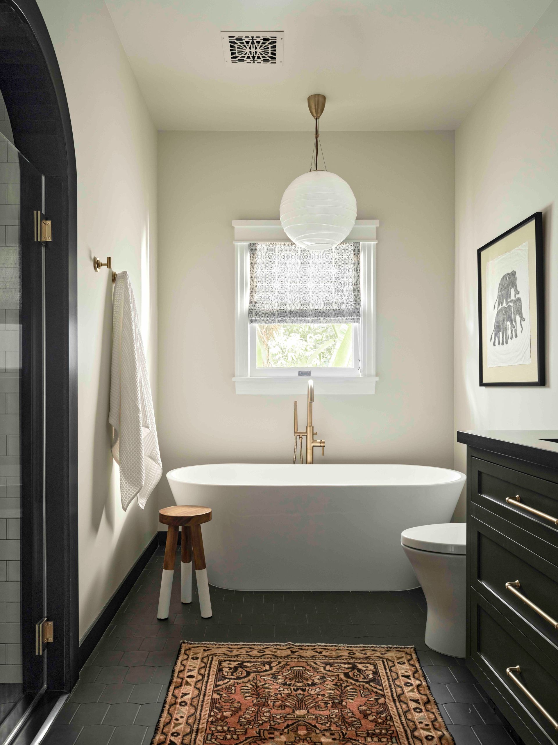 A minimalist bathroom featuring a white soaking tub, dark cabinetry, a pendant light, and a patterned rug on a dark floor.