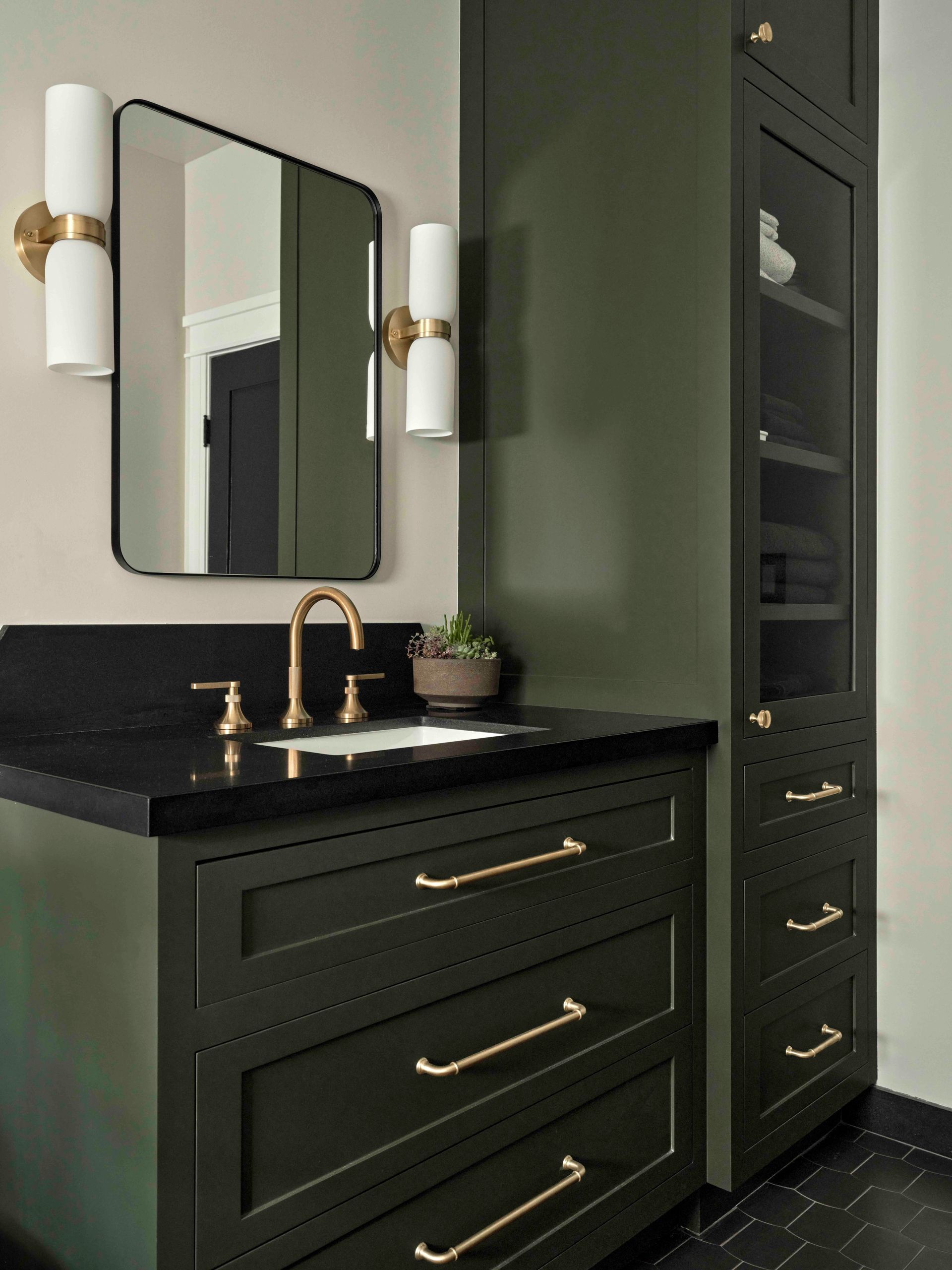 A bathroom vanity with dark green cabinets, a black countertop, gold hardware, and a modern wall-mounted mirror.