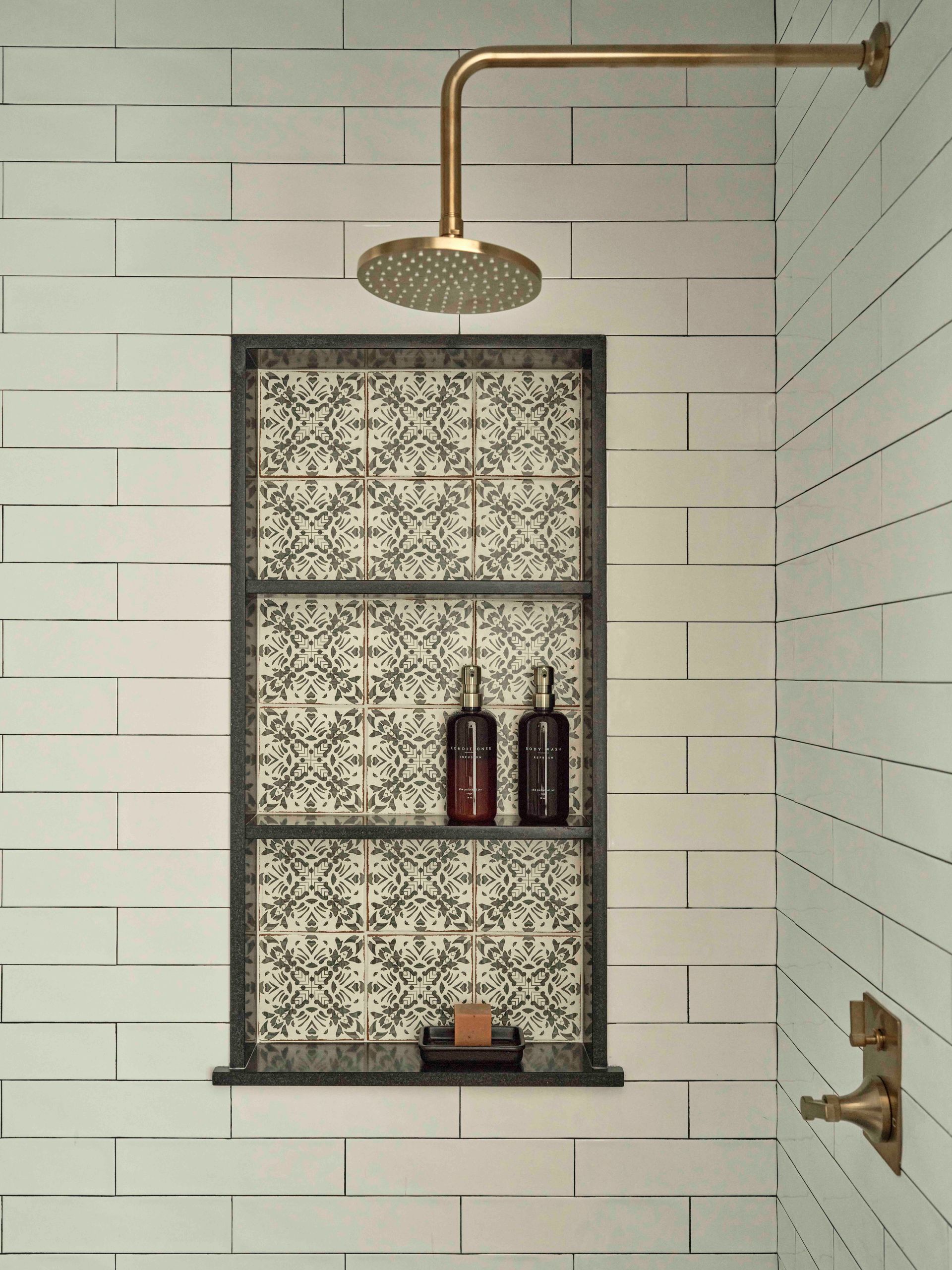A shower with white subway tile, a recessed niche featuring patterned tiles and soap bottles, and brass fixtures.