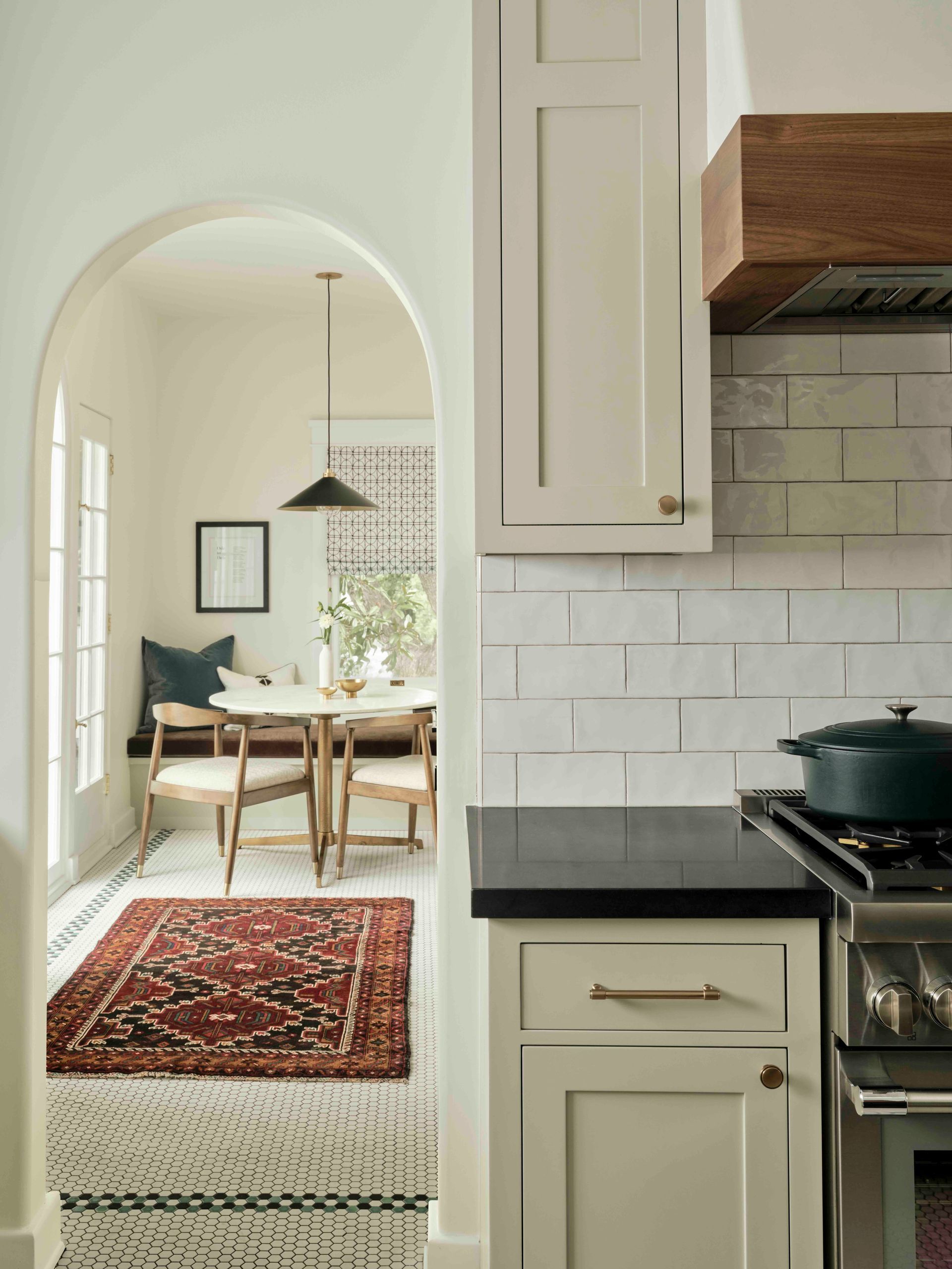 A view through an arched doorway from a light-colored kitchen into a small dining area with a patterned rug and bench.