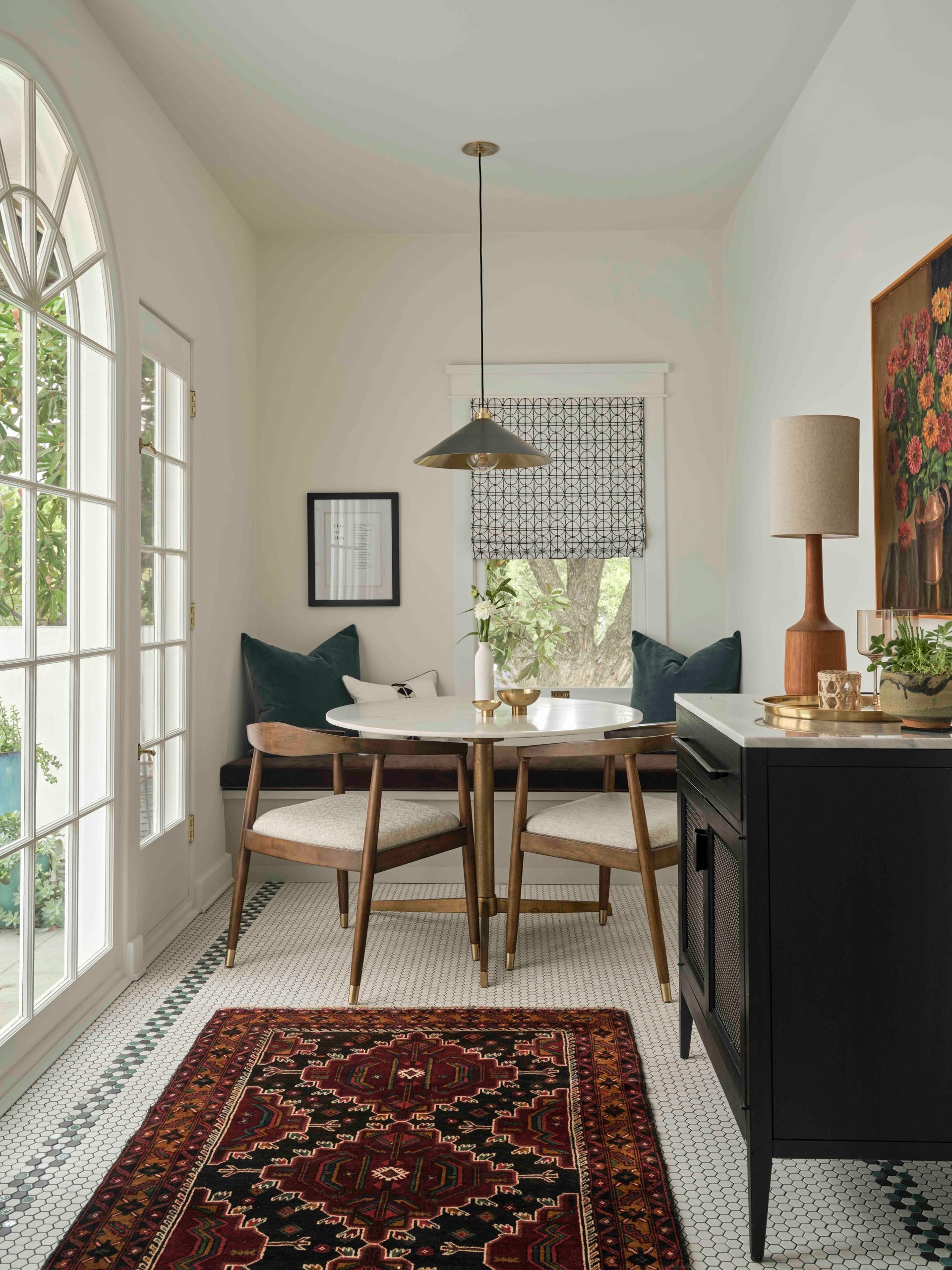 A bright dining nook features a round table with two wooden chairs, a corner bench, a patterned rug, and a pendant light.