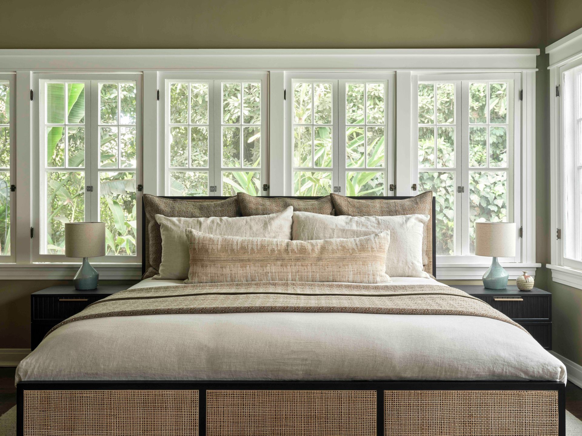A bed with a woven frame sits before a wall of windows, styled with neutral bedding and two small lamps on side tables.