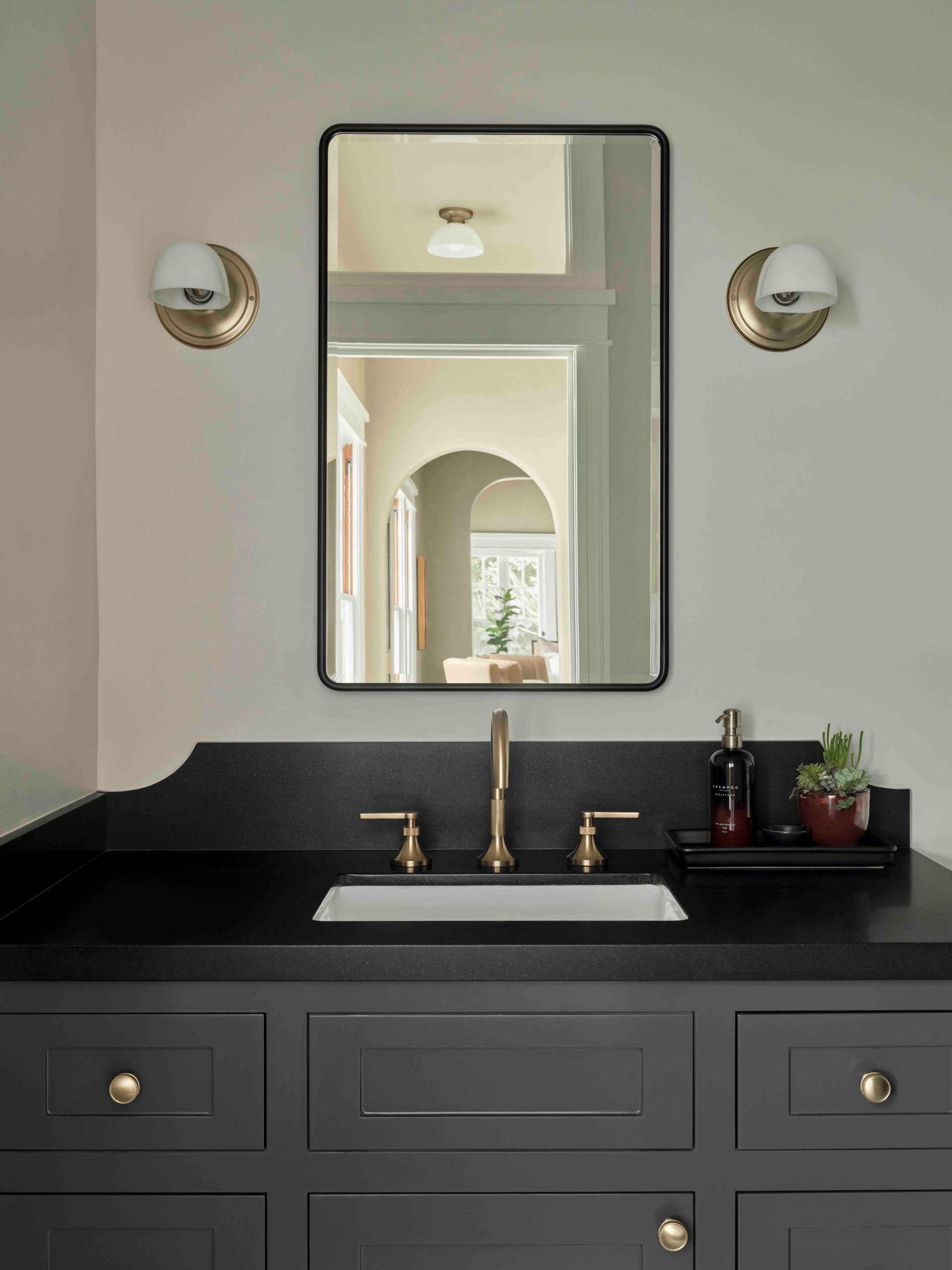 A bathroom vanity with a dark gray cabinet, black countertop, rectangular mirror, and brass fixtures on a light wall.