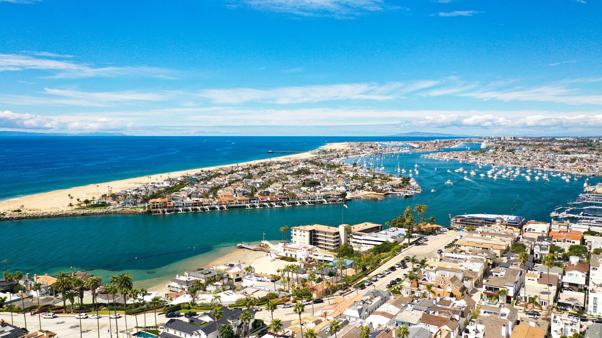 Aerial view of Newport Harbor, California, showing a sandy beach, boat-filled water, and coastal town buildings.