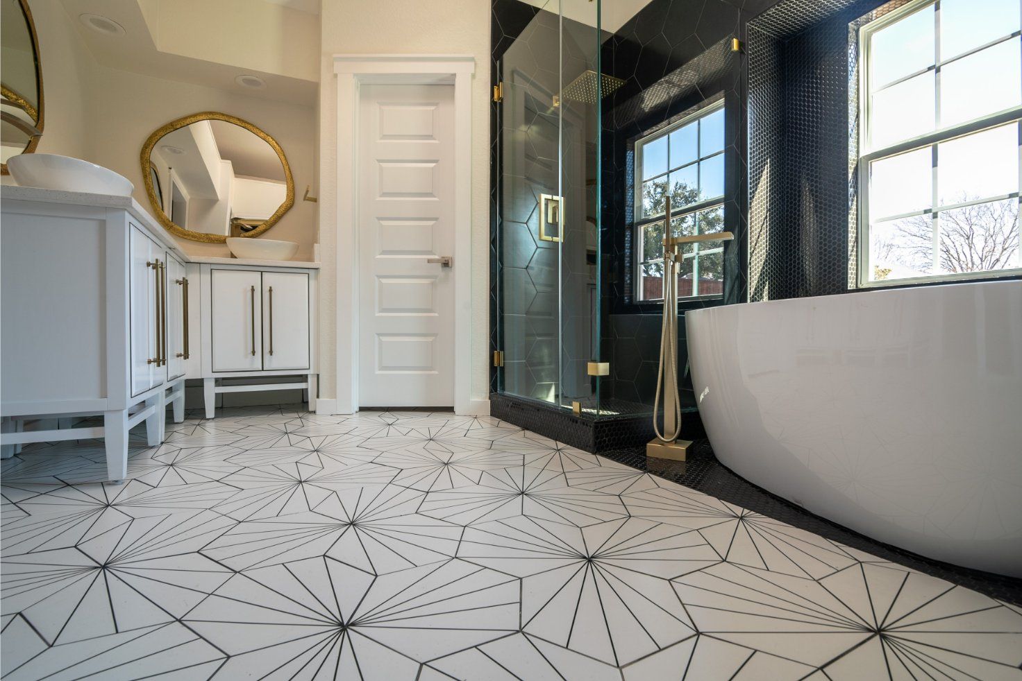 Bathroom with geometric tile floor, white vanity, gold mirror, black tiled wall, glass shower, and freestanding tub.