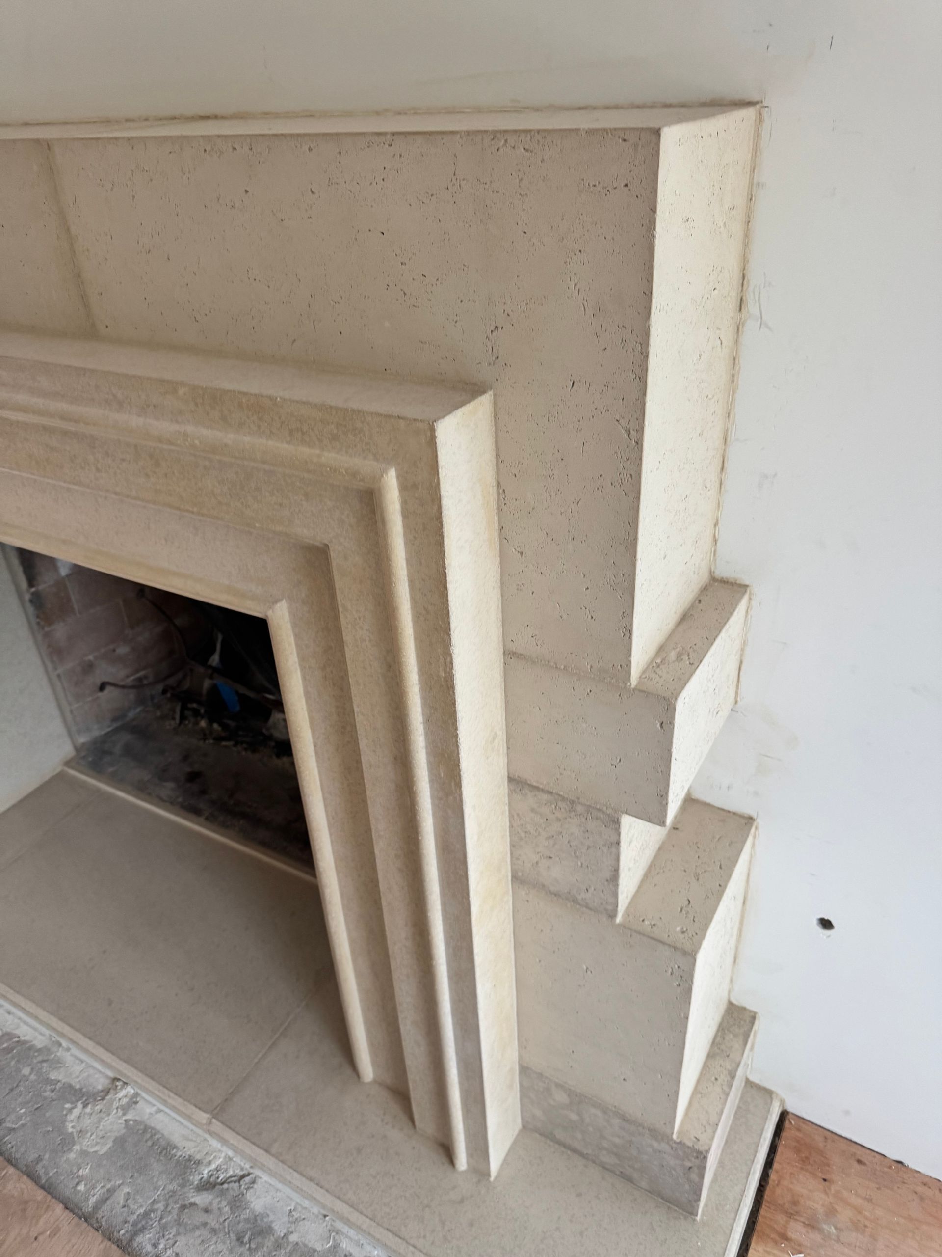 A beige, tiered stone fireplace surround featuring a stepped geometric design, partially installed against a white wall.