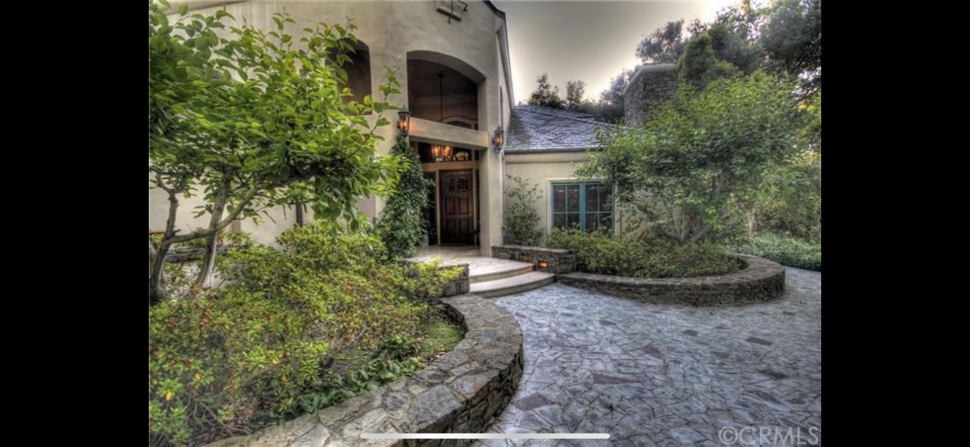 A stone-paved walkway leads to the entrance of a light-colored house framed by lush greenery and curved stone planters.