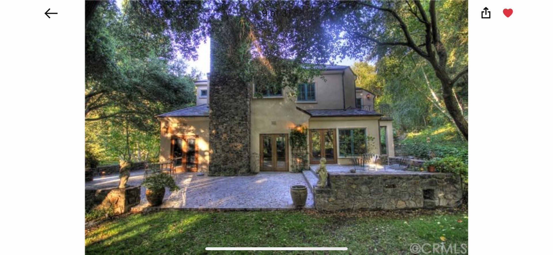 A multi-story house with a prominent stone chimney, patio, and garden, viewed from a lawn surrounded by mature trees.