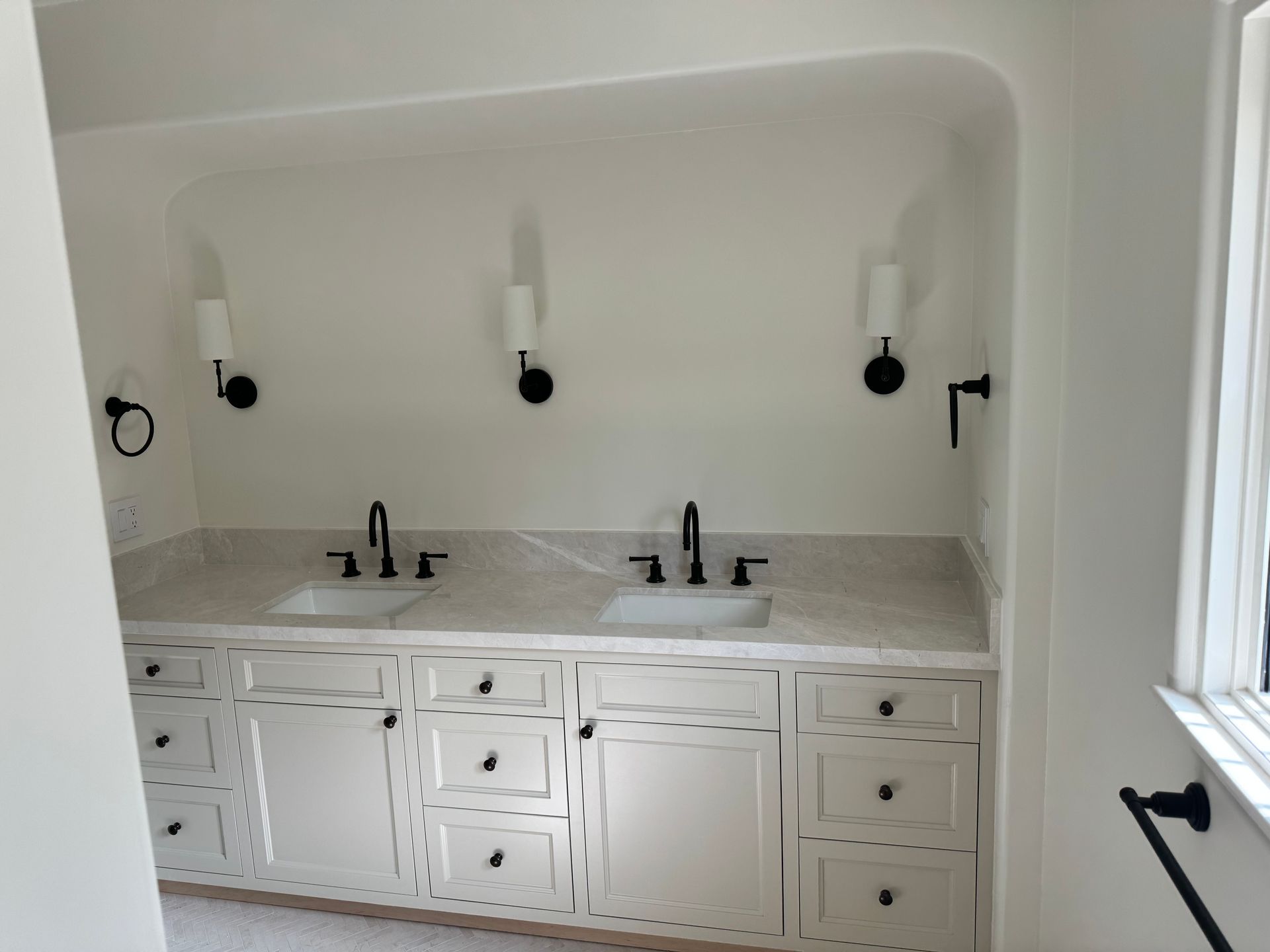 A white double-sink bathroom vanity with black hardware and sconces in a recessed, arched nook.