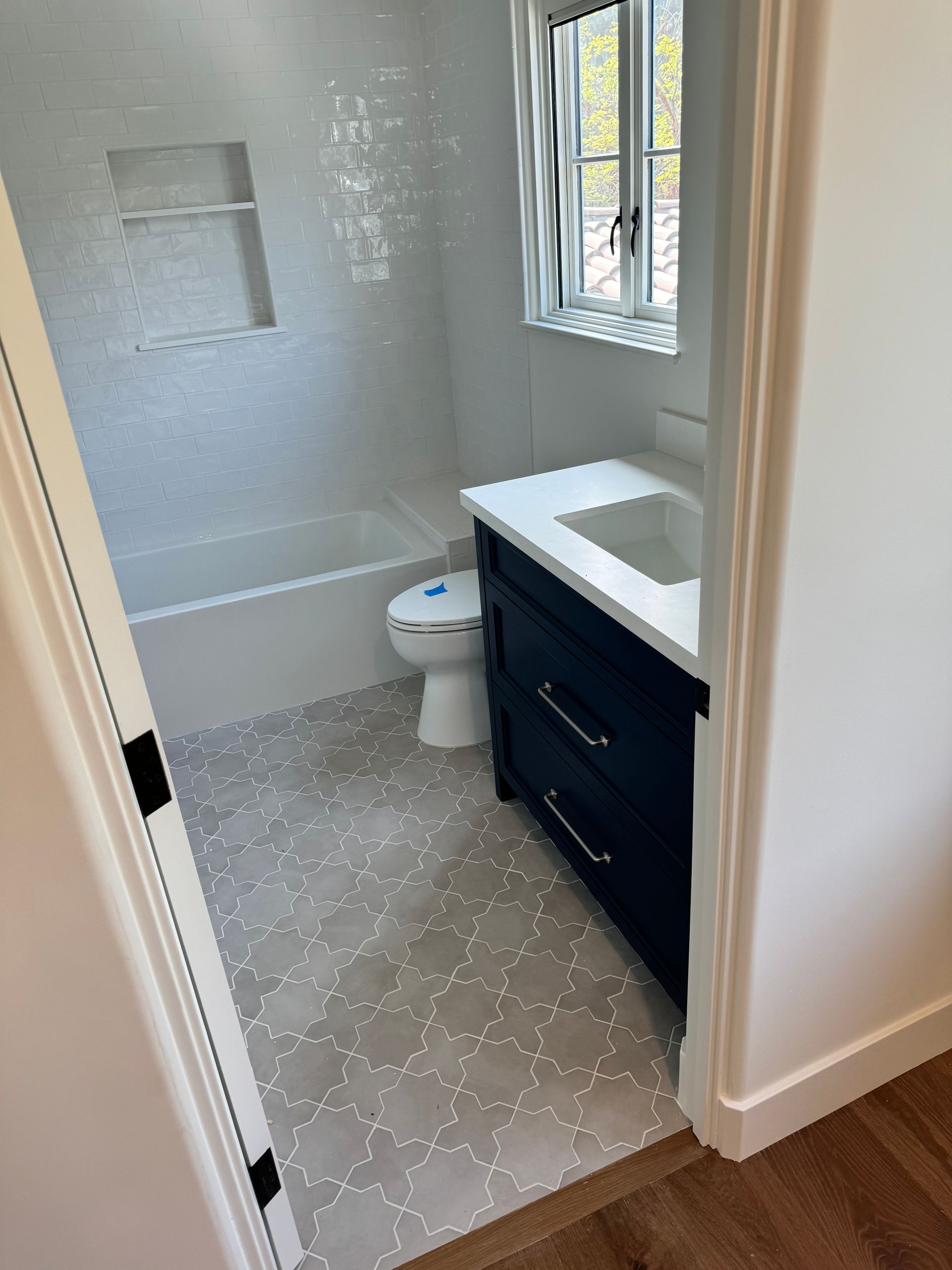A small, modern bathroom with a navy vanity, white countertop, bathtub with a recessed shelf, and light patterned flooring.