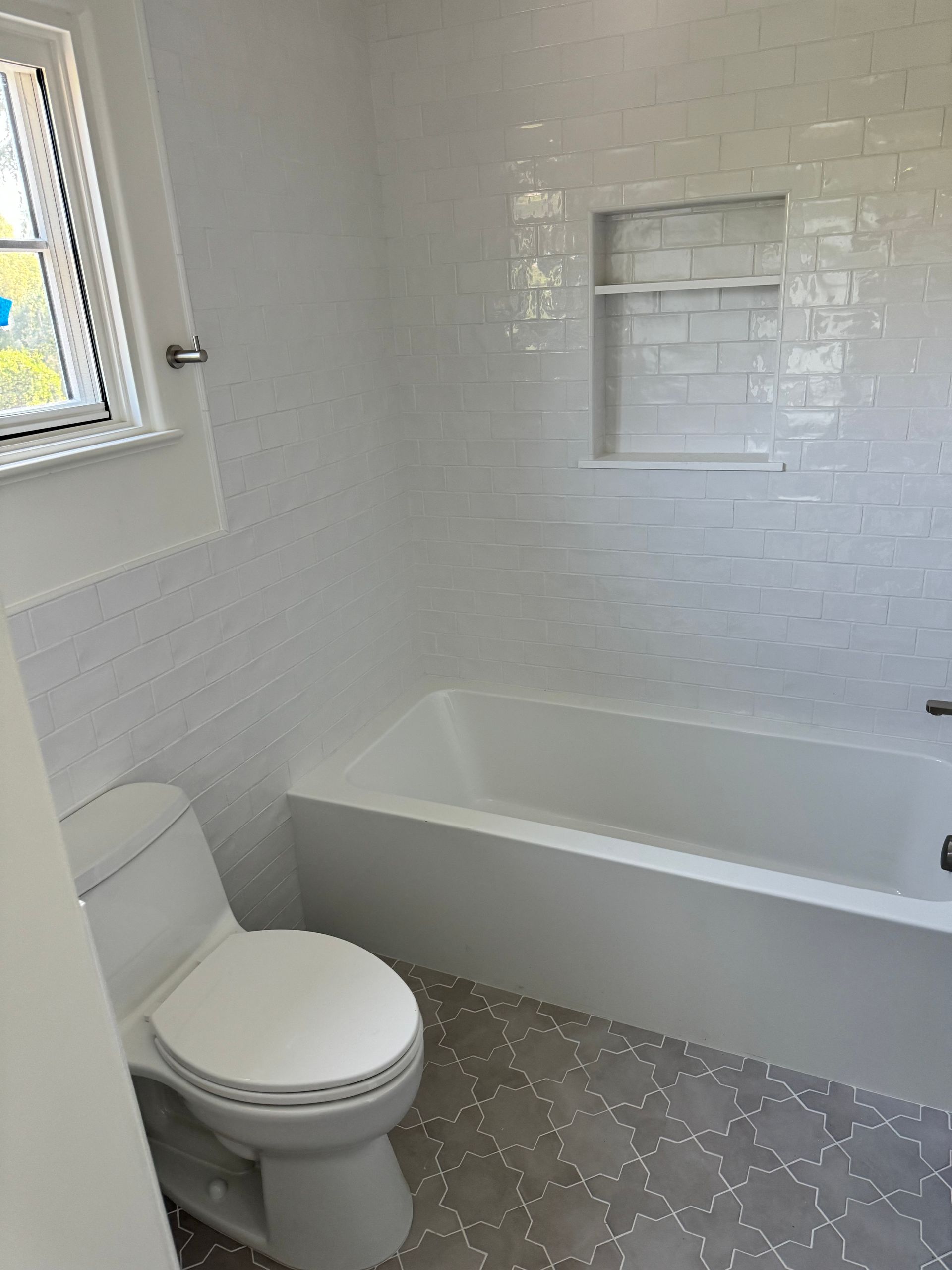 A bright white bathroom featuring a bathtub with an inset tile niche, a white toilet, and patterned gray floor tile.