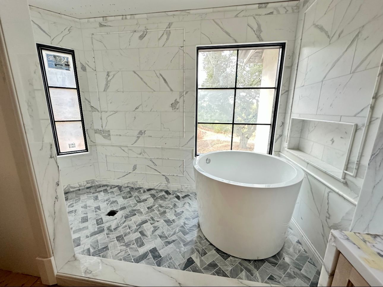 A modern bathroom features white marble tile walls, a patterned gray tile floor, a soaking tub, and two large windows.