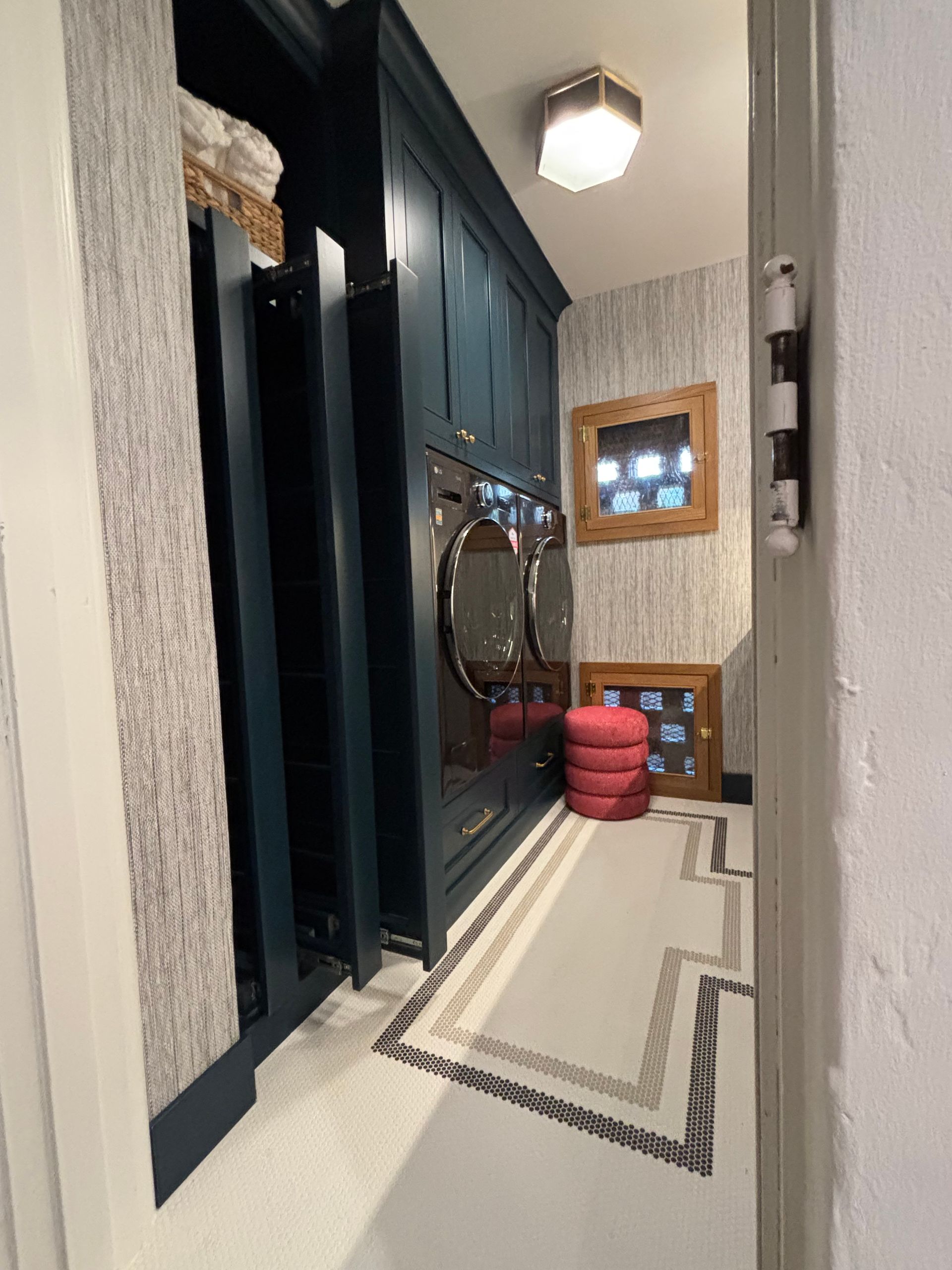 A laundry room with dark blue cabinetry, a patterned rug, light textured wallpaper, and a stack of red cushions.