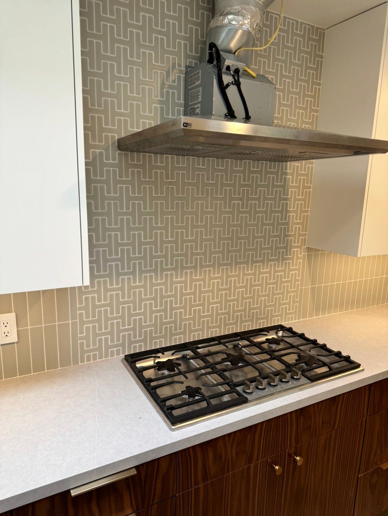 A modern kitchen featuring a stainless steel range hood over a gas cooktop, with patterned tile backsplash and wood cabinets.