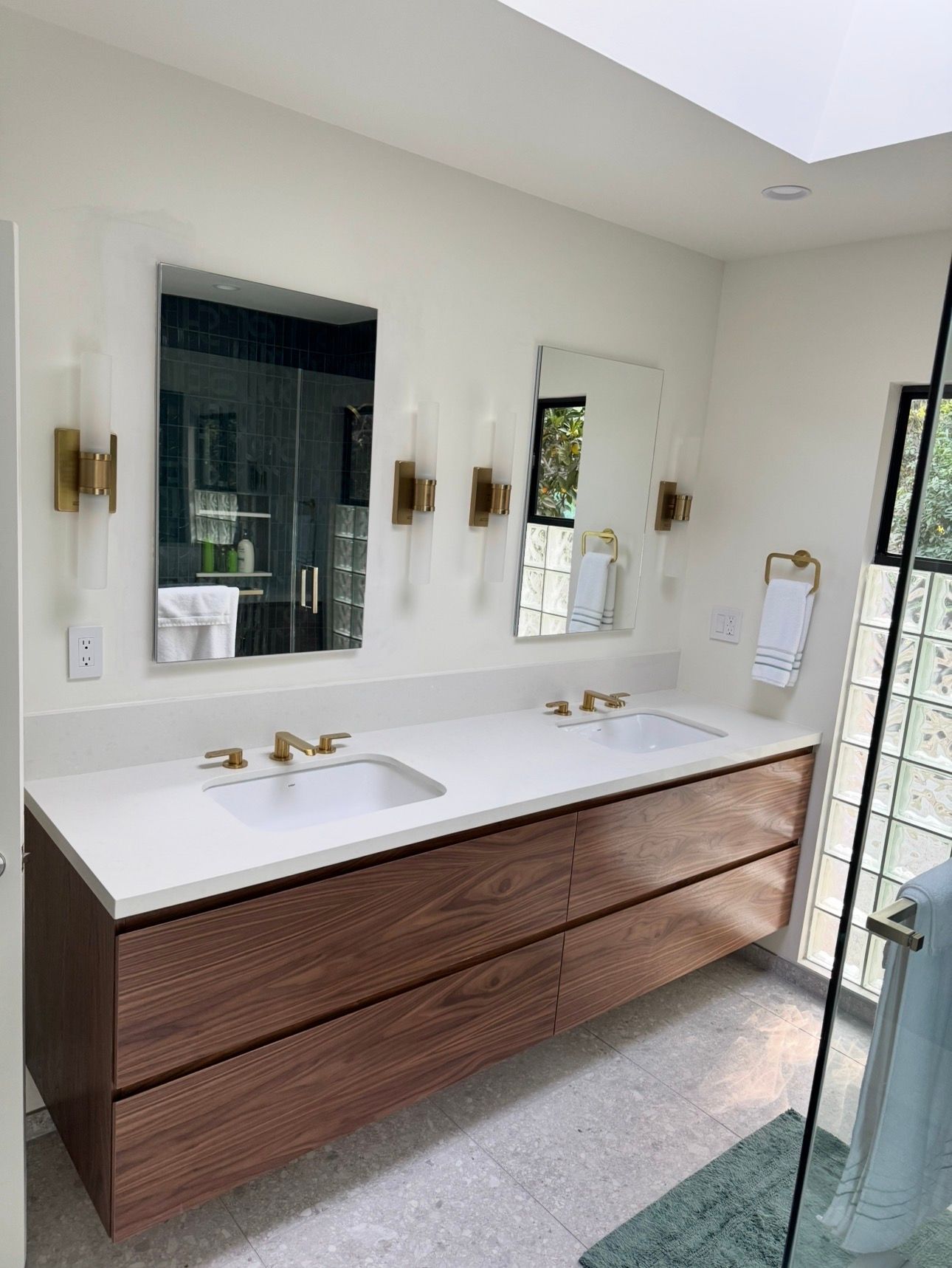 Modern double-sink bathroom vanity with wood drawers, white countertop, gold fixtures, two mirrors, and wall sconces.