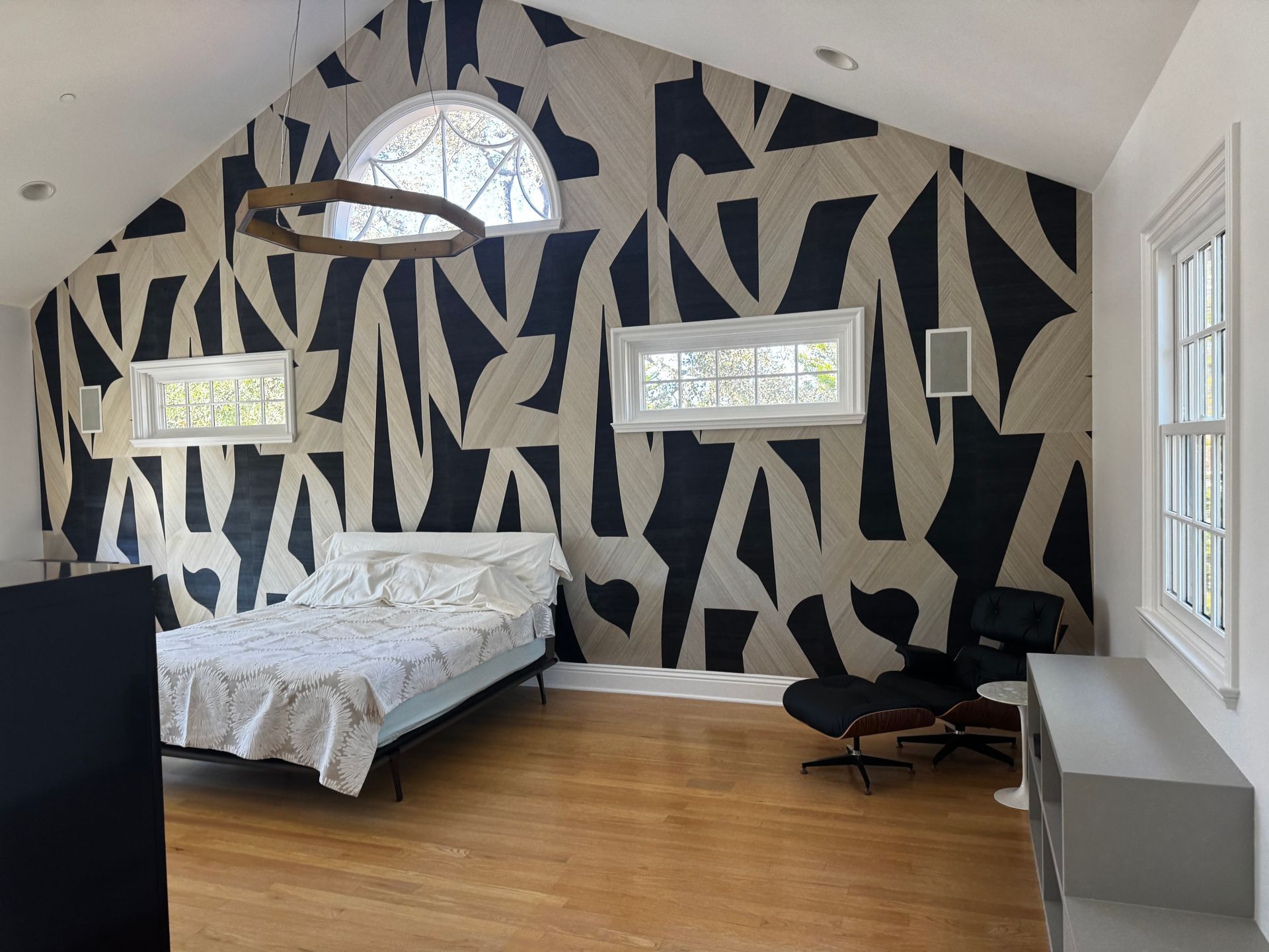 A bedroom with a bed and lounge chair against a large wall featuring a bold, black-and-tan abstract geometric pattern.