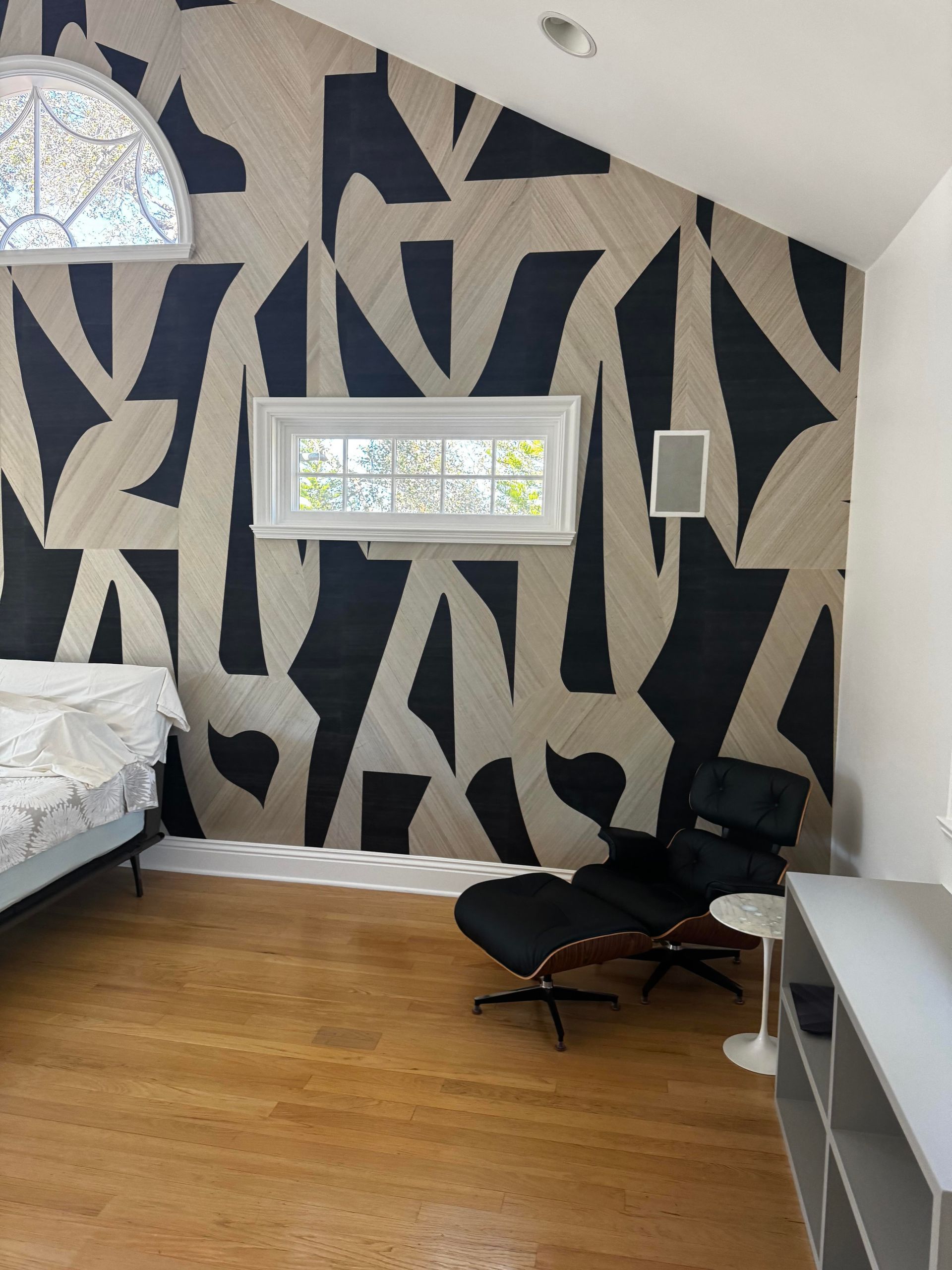 A bedroom corner with a bold, black-and-tan abstract patterned wall, a black lounge chair, and a small side table.