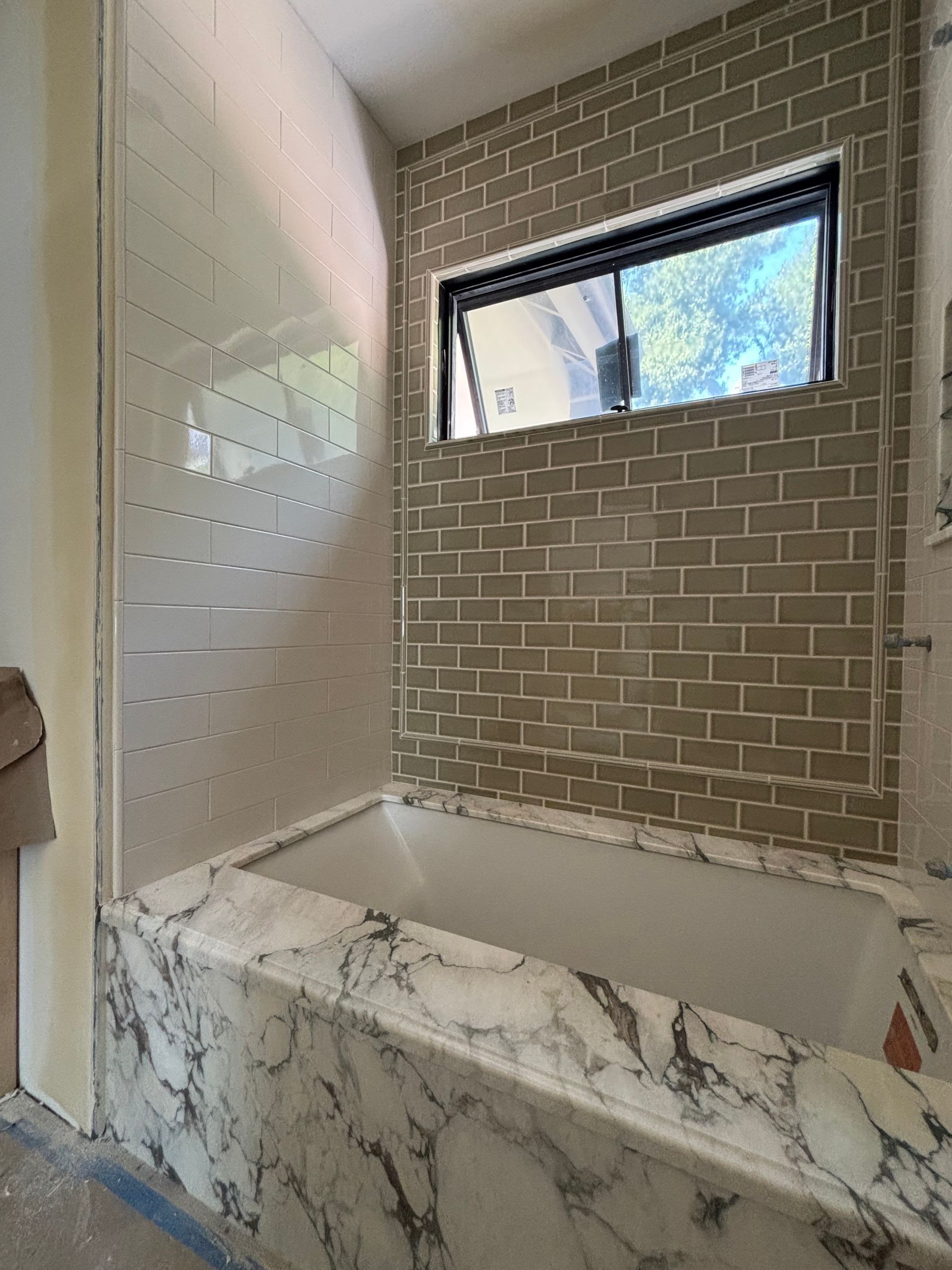 Bathtub with marble-patterned surround, tiled walls, and a window in a bathroom under construction.