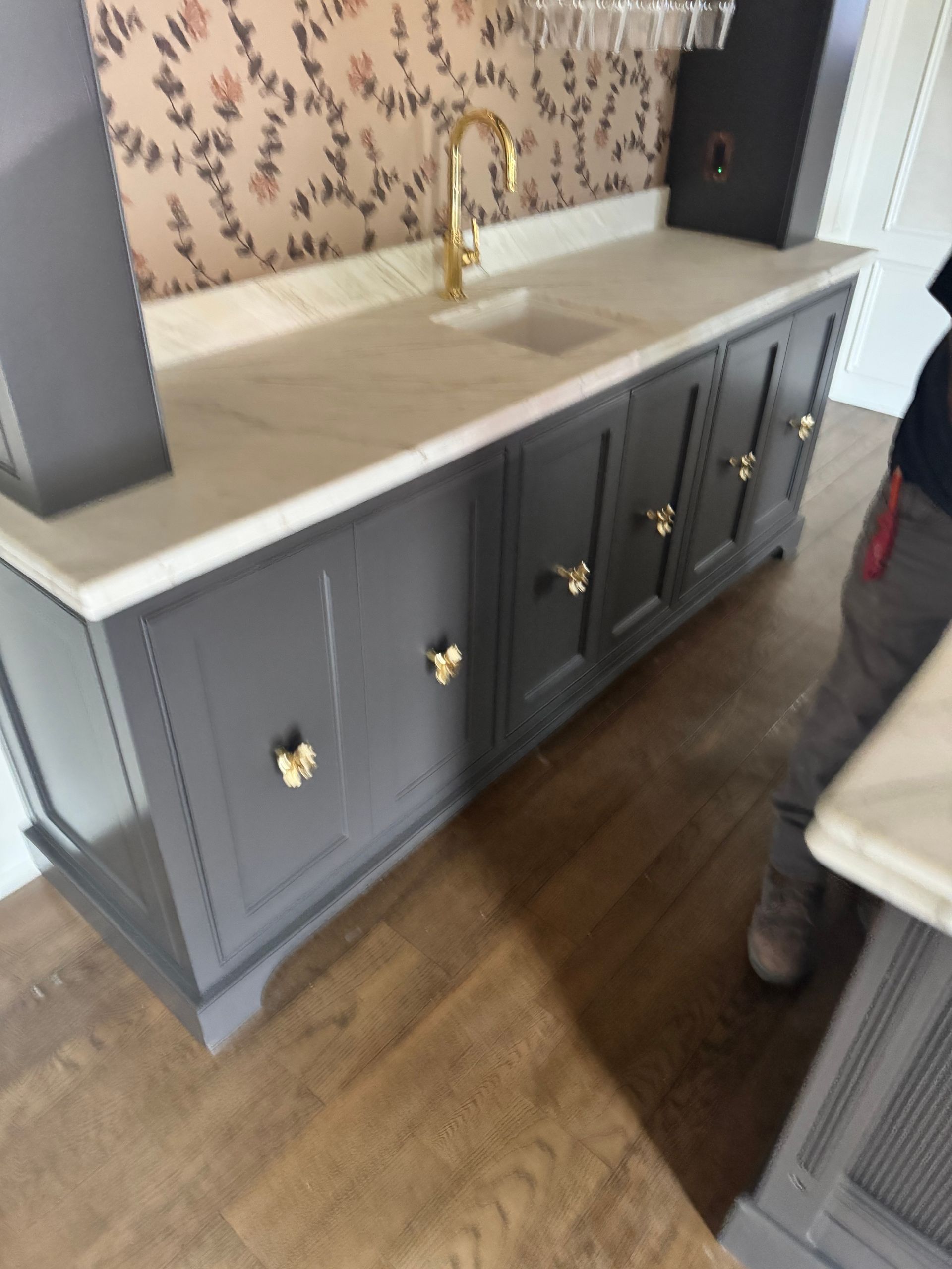 A dark grey wet bar with brass hardware, light stone countertop, and floral wallpaper, with a person standing to the right.