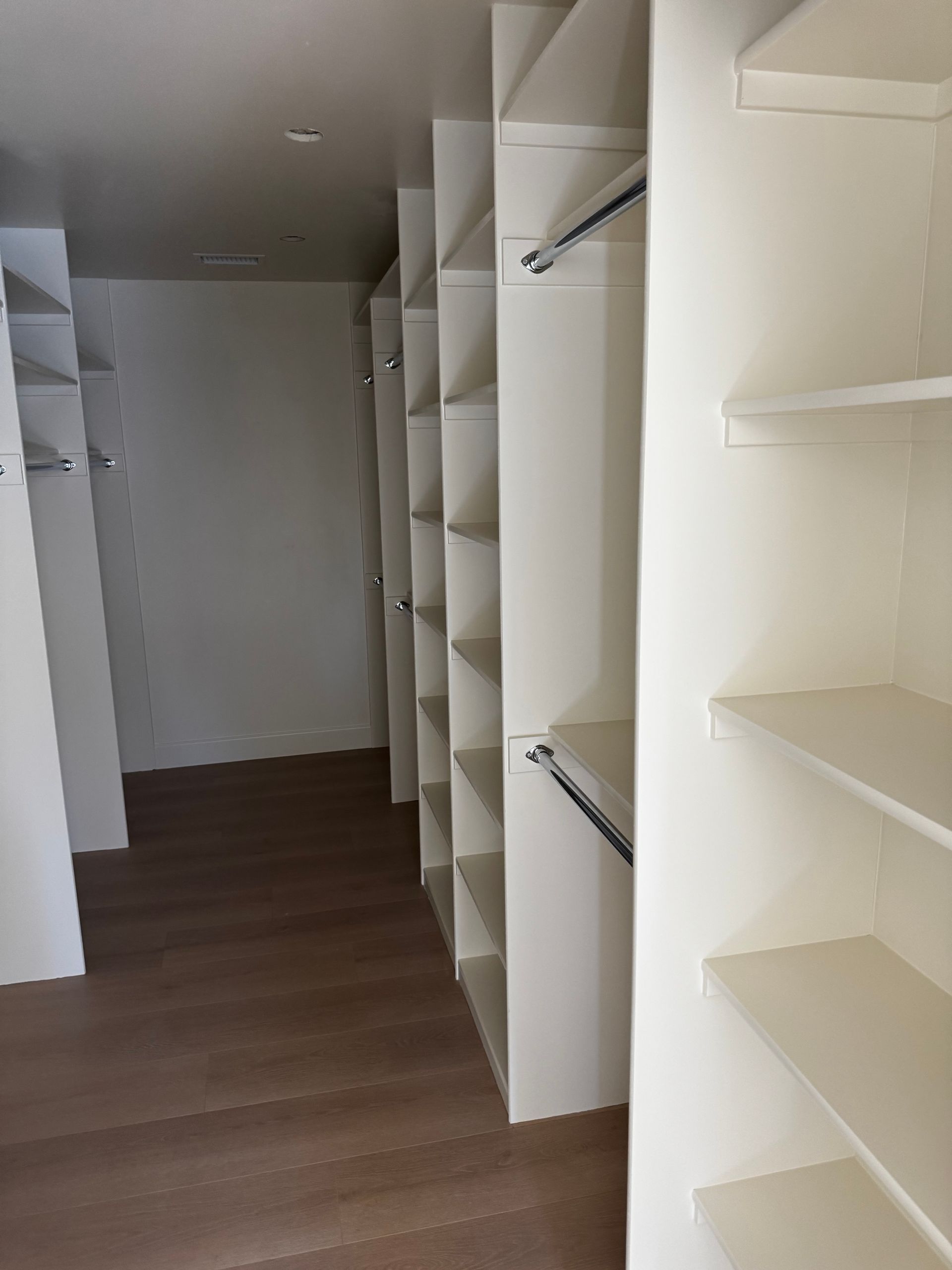 A walk-in closet featuring white shelving units, hanging rods, and wood-look flooring.