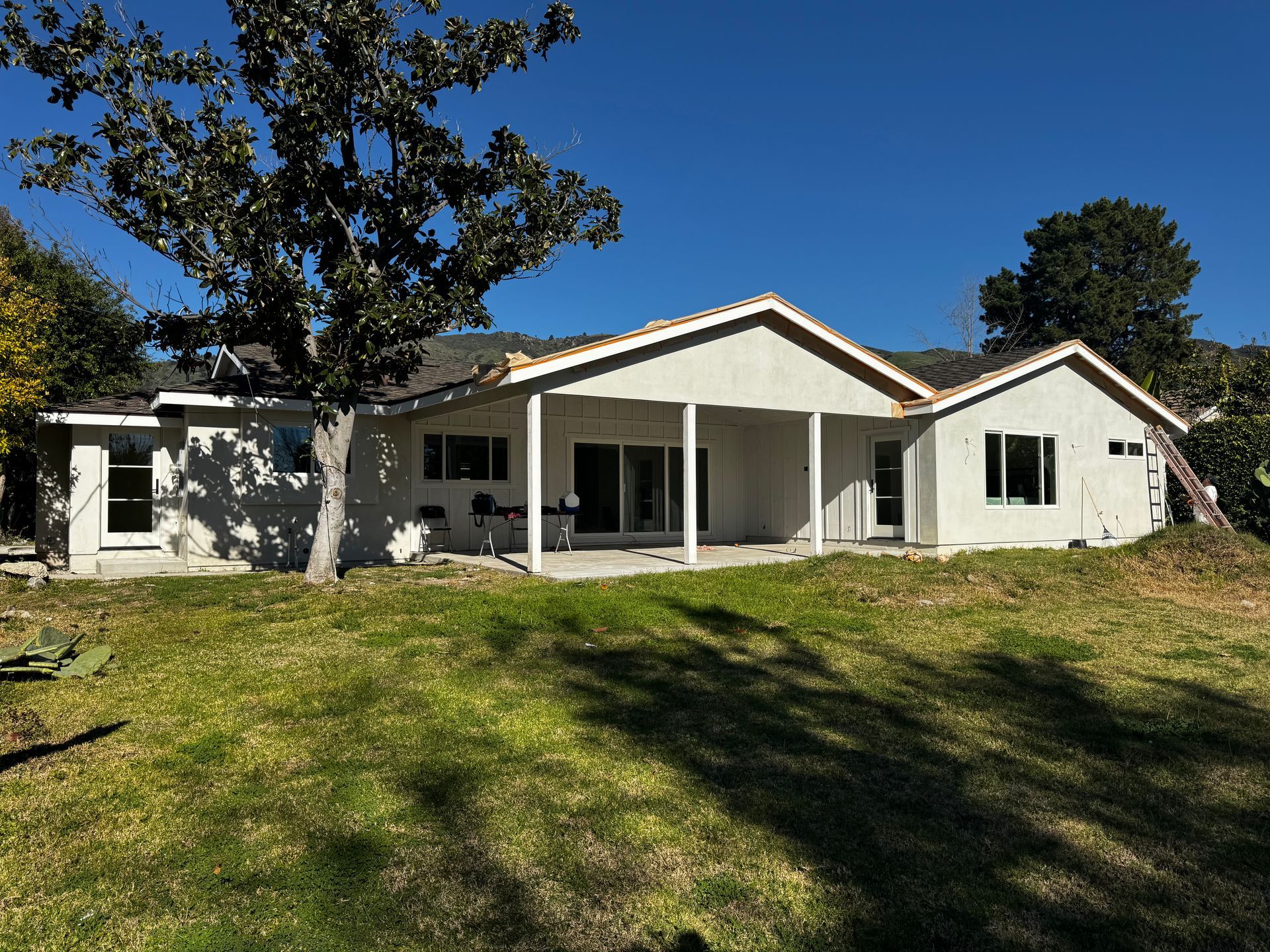 A light-colored, single-story house with a covered patio under construction, surrounded by a lawn and trees under a blue sky.