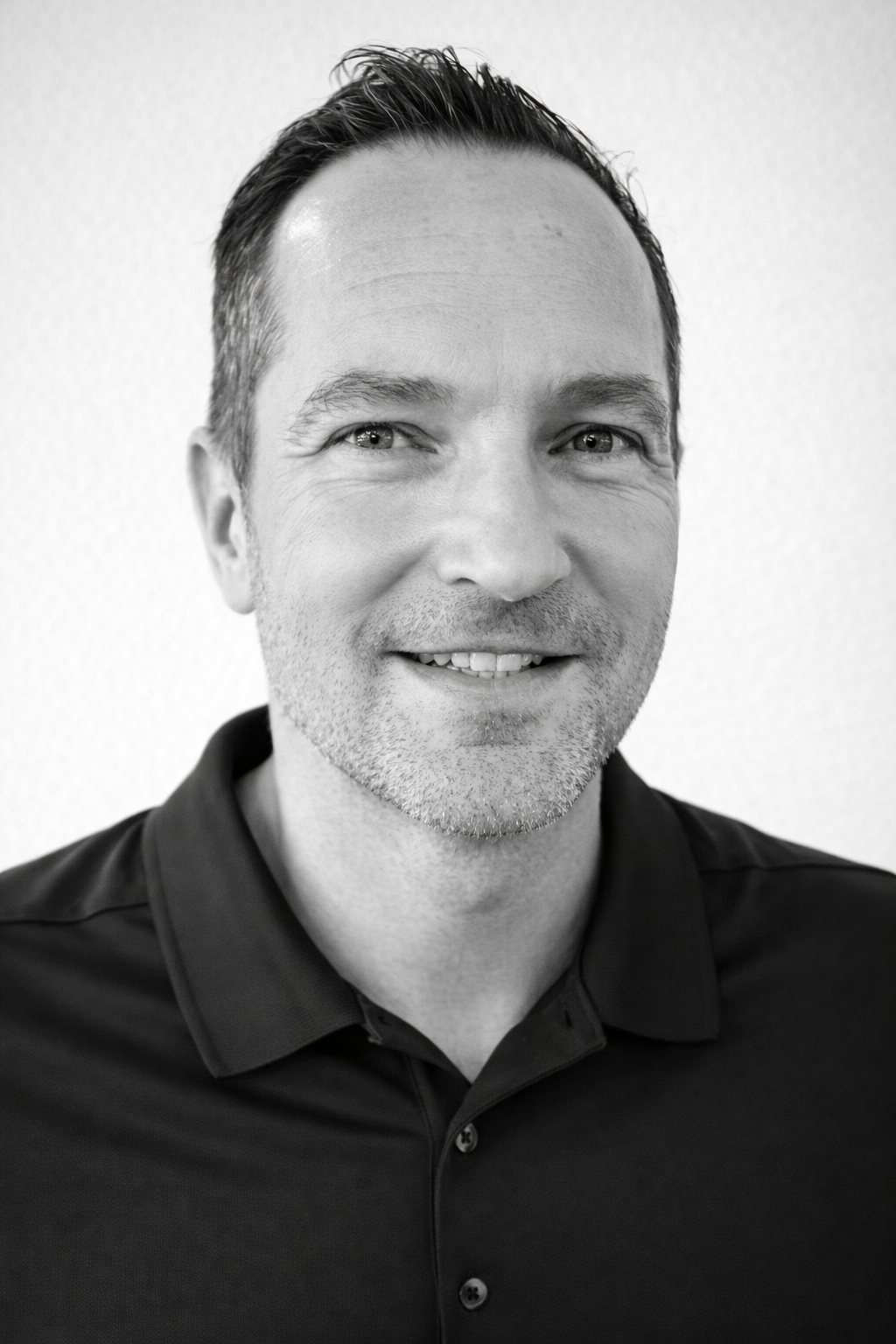 Headshot of a person with short hair, wearing a dark polo shirt against a white background, smiling at the camera.