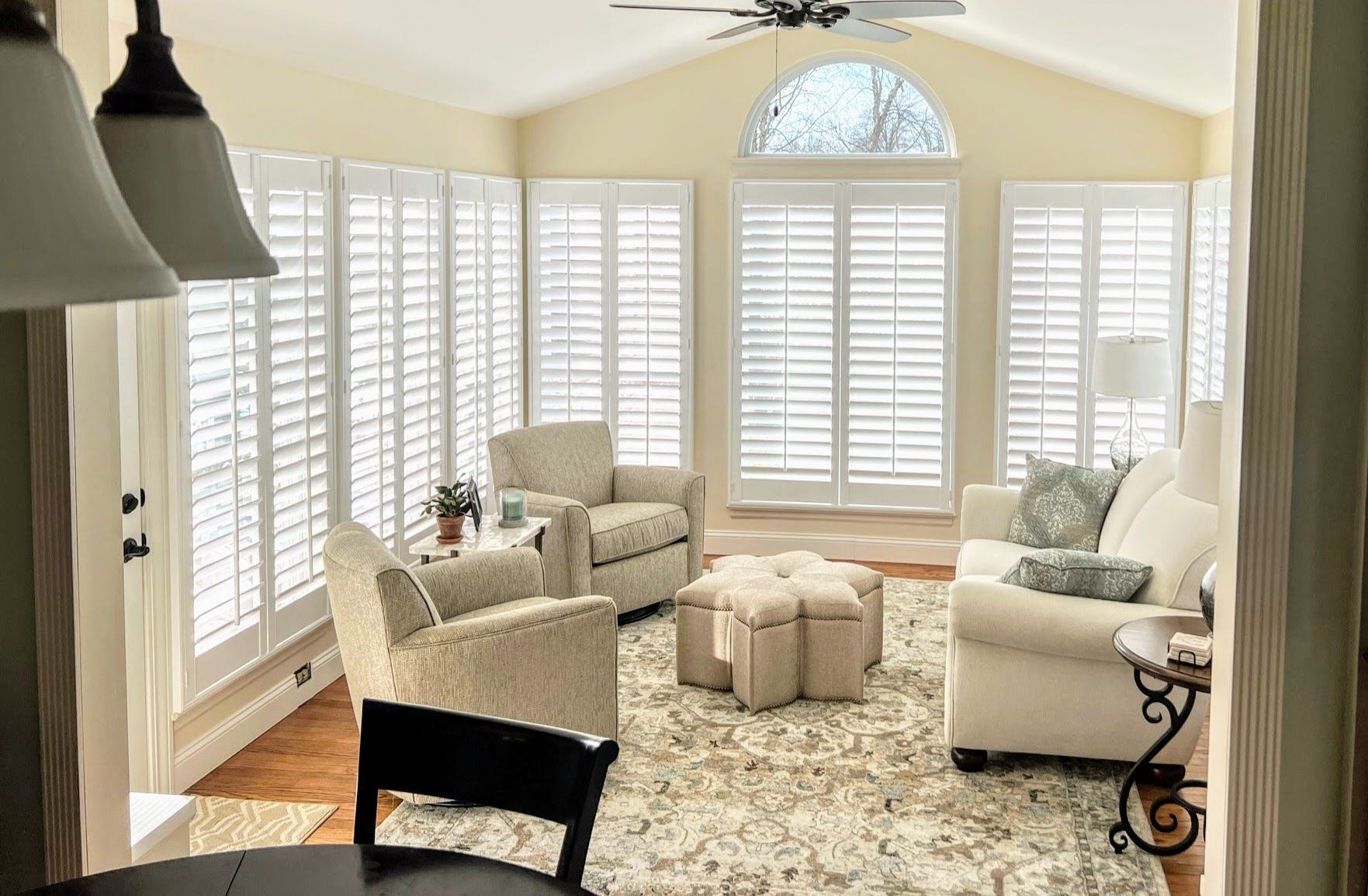 Sunroom with white shutters, cream-colored furniture, and a neutral rug. Sunlight streams through the windows.