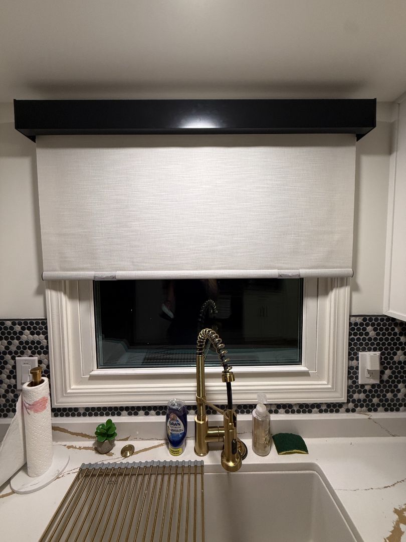 Kitchen window with partially lowered cream-colored roller shade, black frame. Gold faucet, white sink, patterned backsplash.