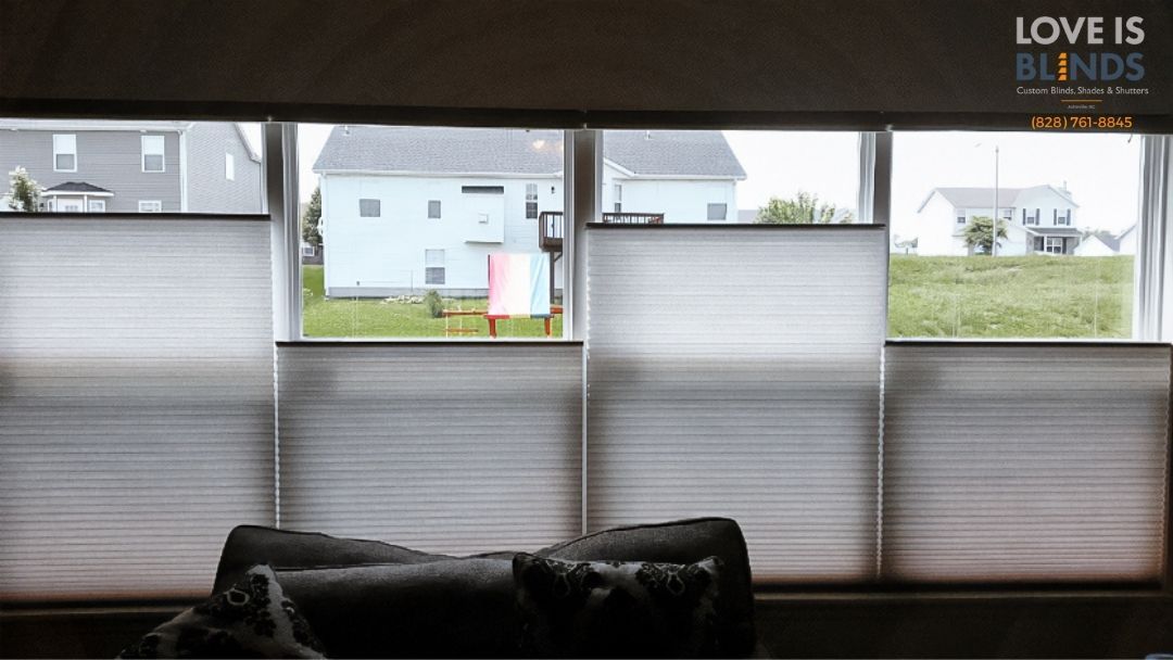 Closed blinds on a window overlooking houses and grass. Soft light filters through.