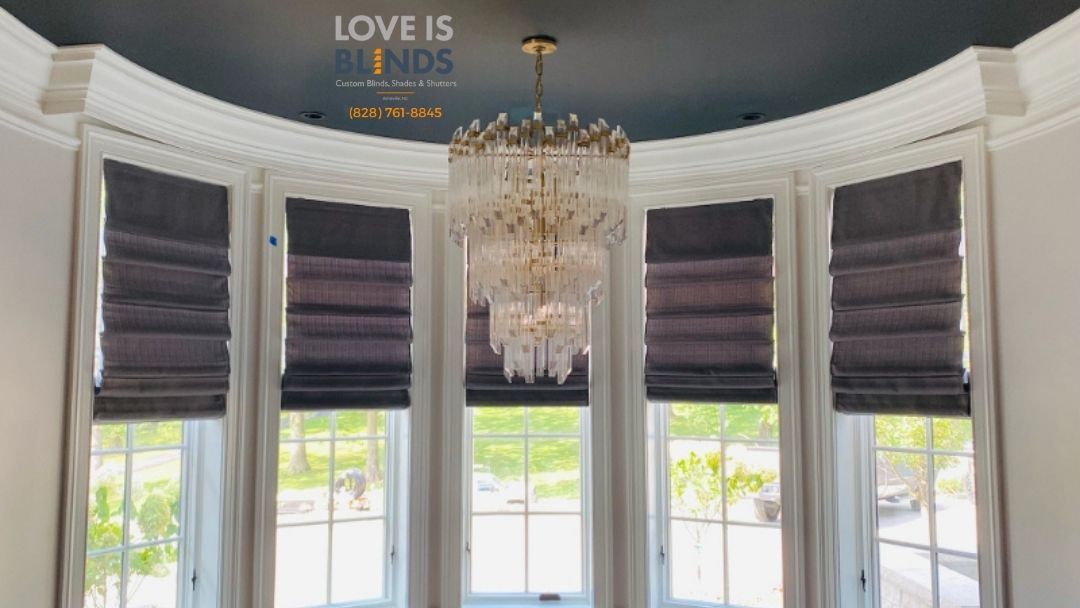 Bay window with dark gray Roman shades, ornate chandelier, and view of greenery.