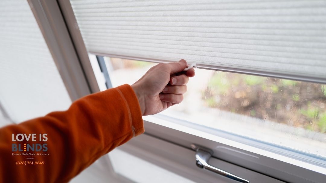 A person's hand pulling a string to lower a pleated white window shade.